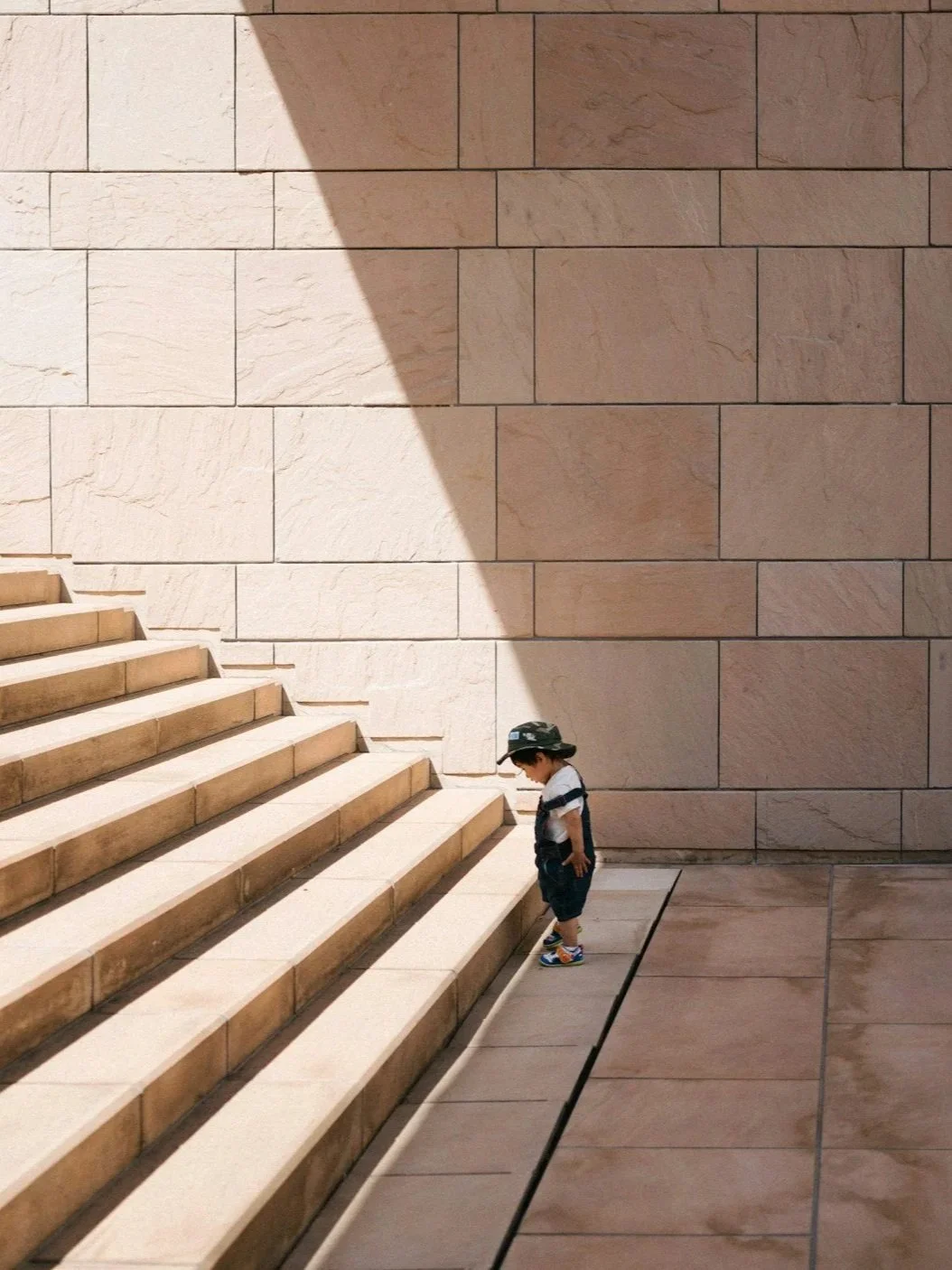 A toddler dressed in overalls and a hat stands at the bottom of sandstone-colored steps. The sun illuminates the steps.