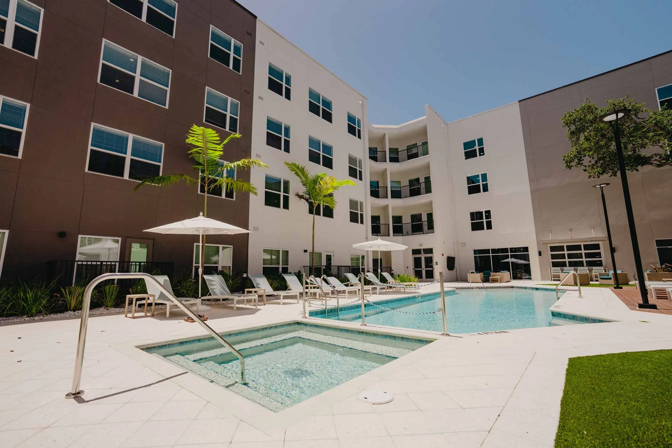 A modern apartment complex courtyard with a swimming pool, sun loungers, umbrellas, trees, and a multi-story building with numerous windows and balconies under a clear blue sky.