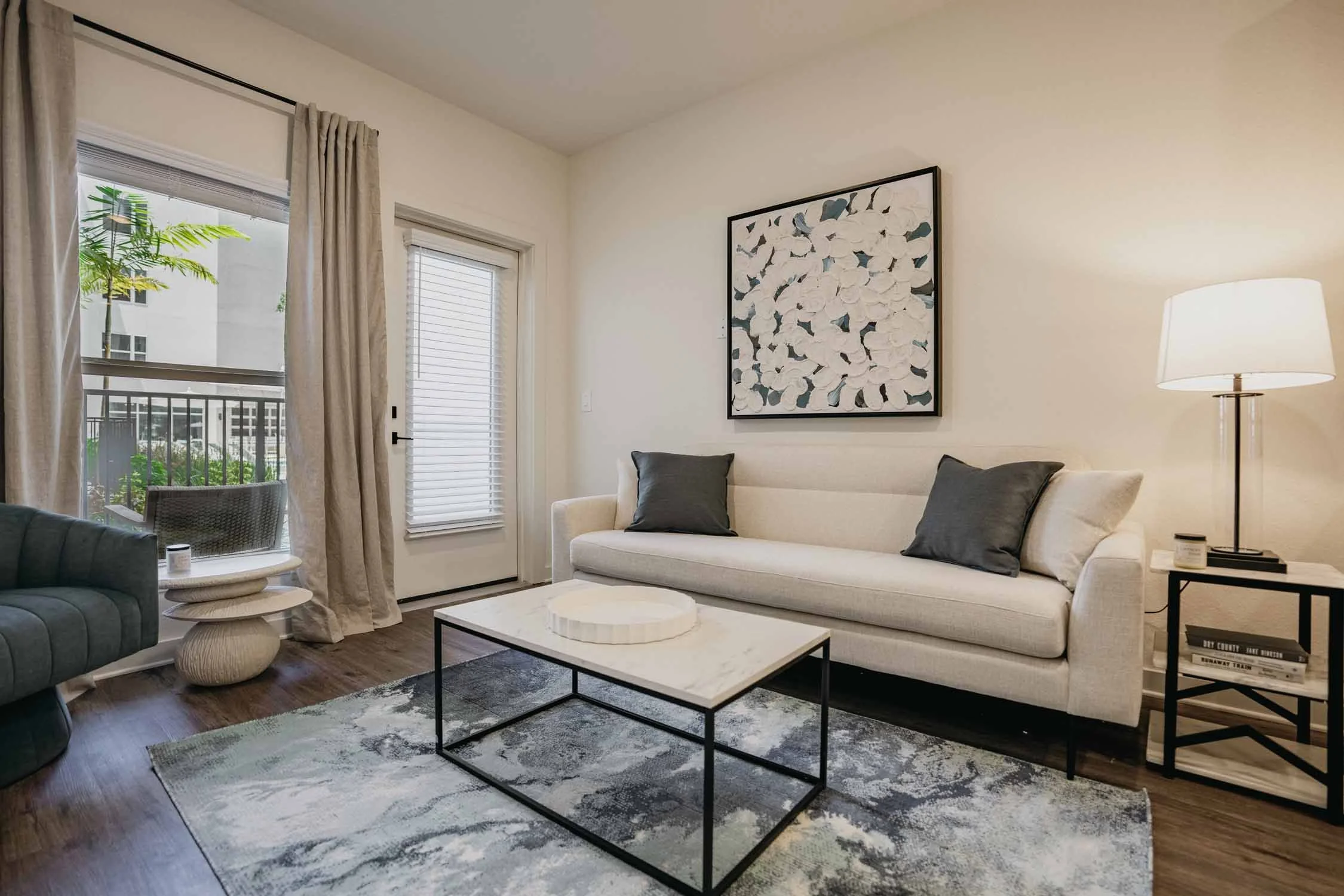 Modern living room with white sofa, black cushions, abstract artwork on the wall, glass-top coffee table, and a standing lamp. Two large windows with curtains and blinds, and a sliding door leading to a balcony with outdoor furniture and green plants.