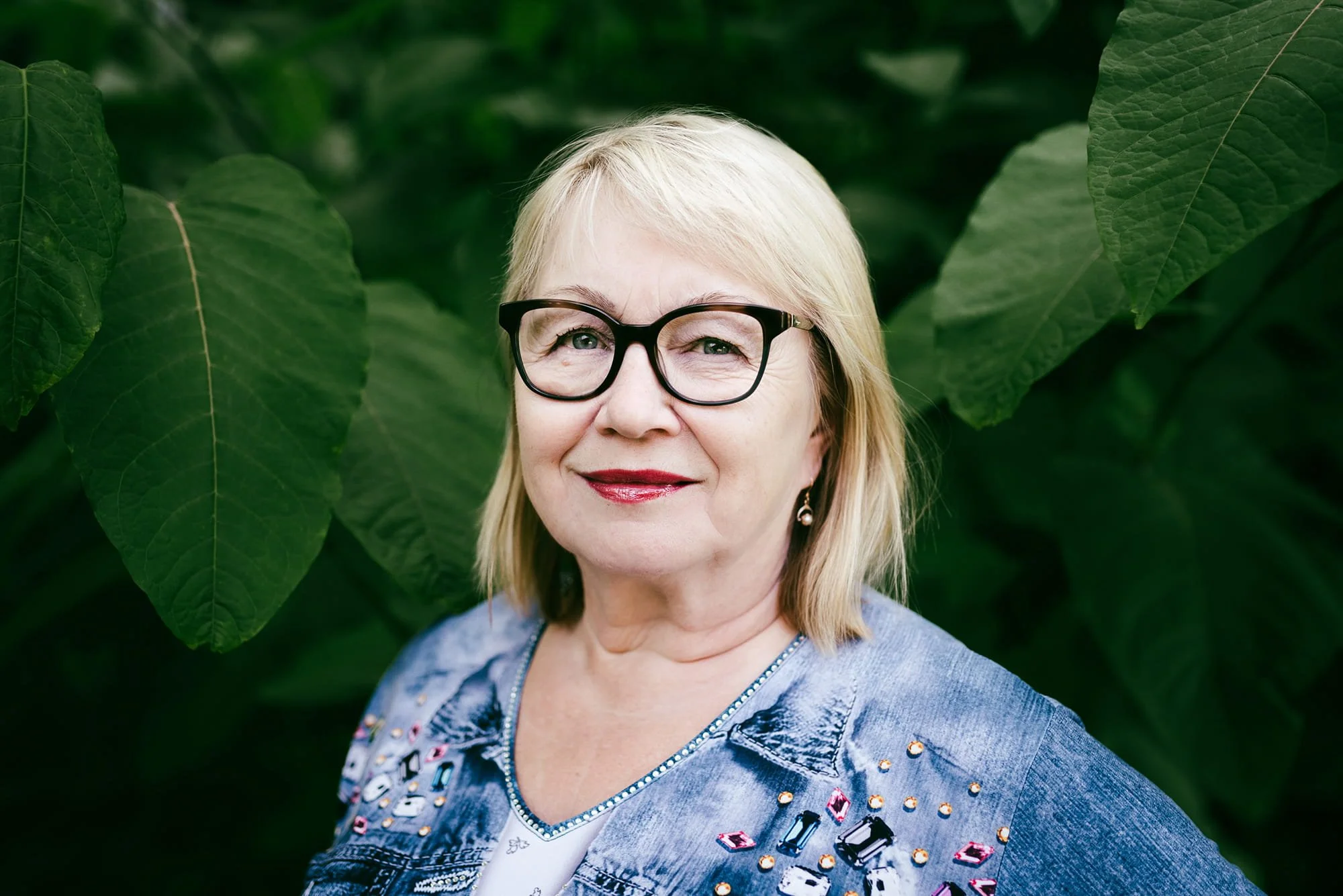 Potrettikuva hymyilevästä iäkkäämmästä naisesta vehreän pensaan edessä