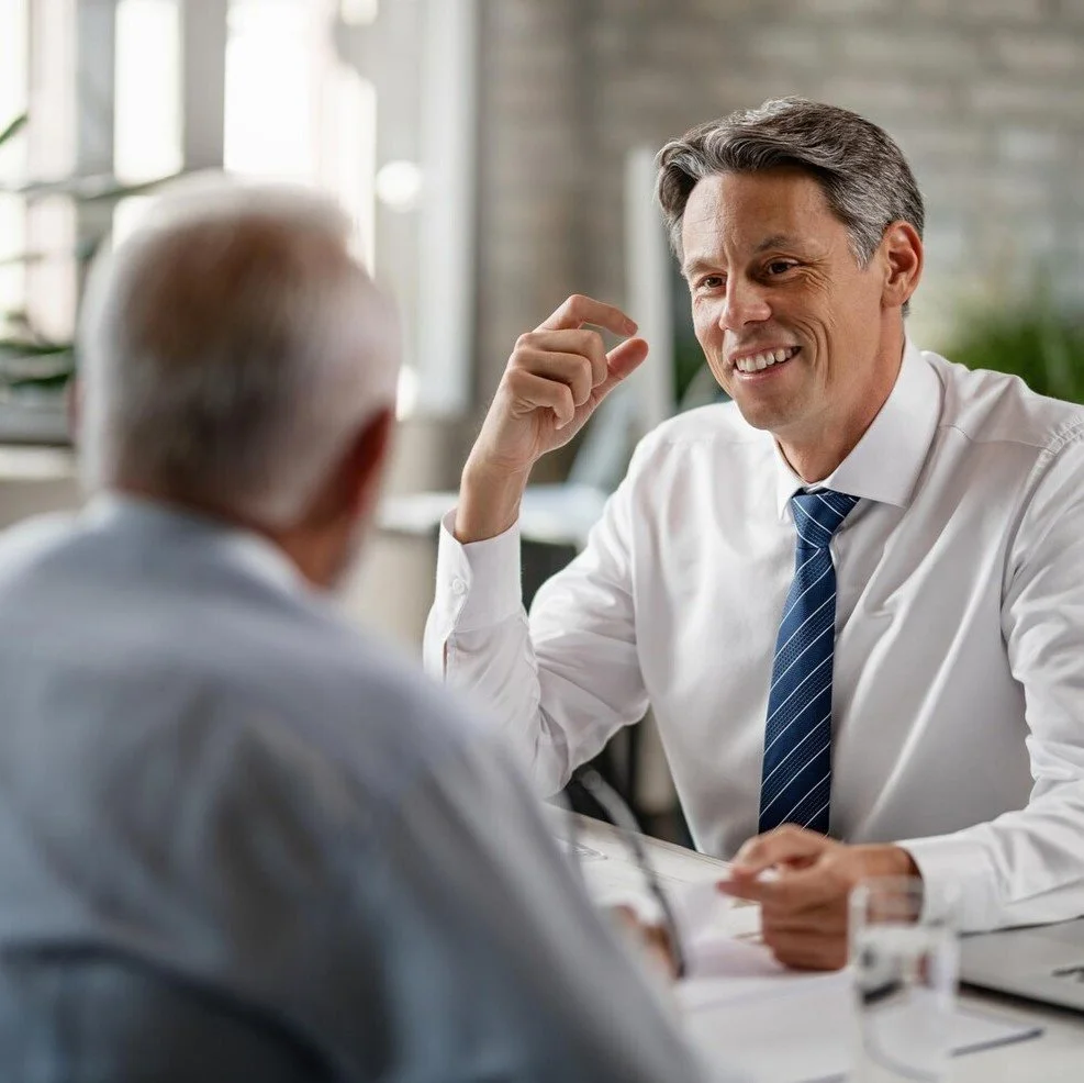 A man in a white shirt and blue tie smiling and talking to an older person in an office setting.