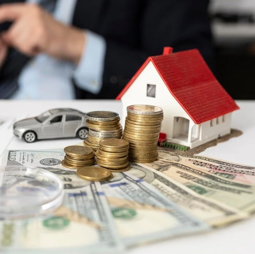 Stacks of coins, a small model house with a red roof, and a toy car on a table, with US dollar bills underneath.