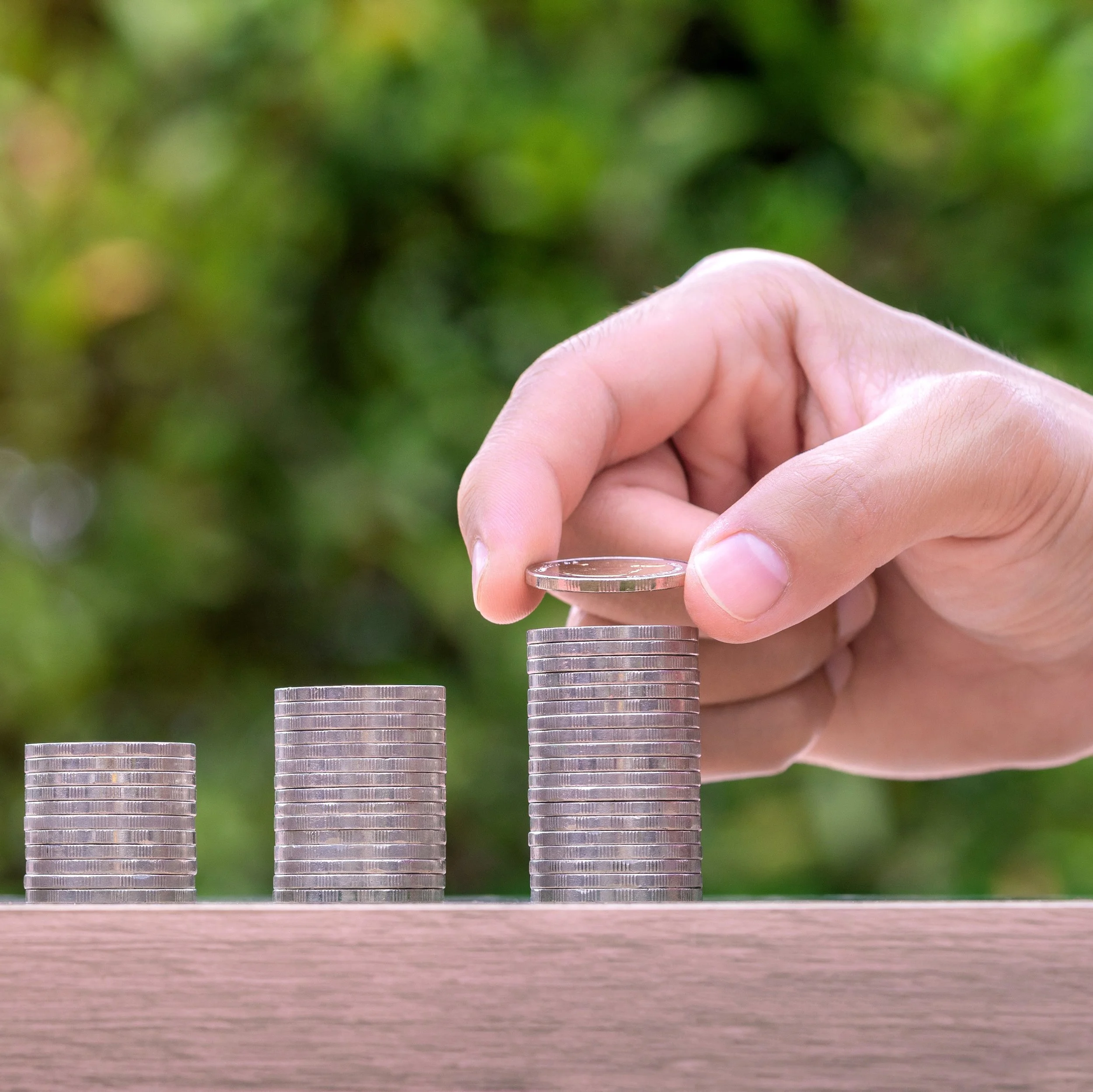 A hand stacking coins on top of each other in increasing height from left to right, with a background of green foliage.