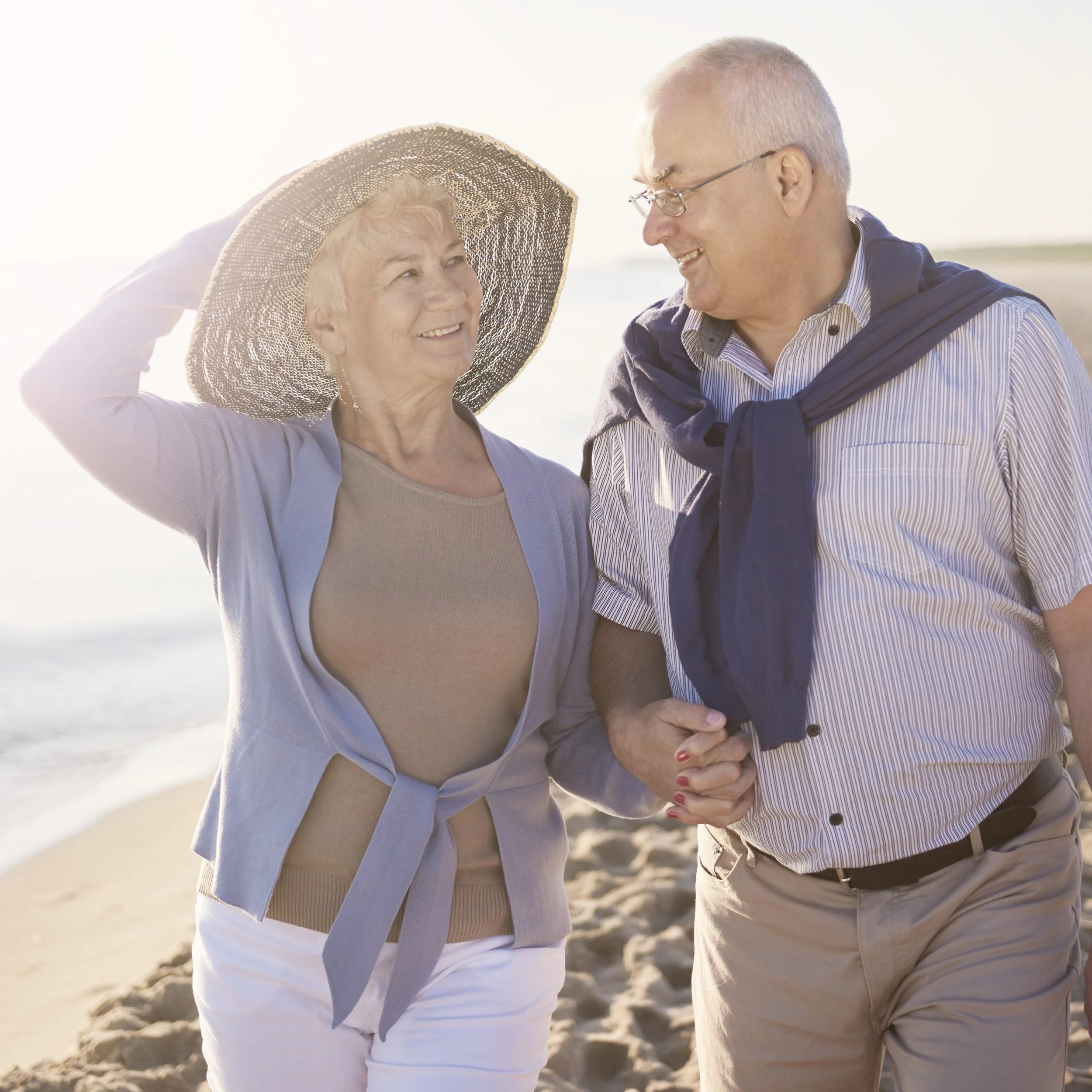 An elderly couple smiling and walking hand in hand on a sandy beach during sunset, enjoying a sunny day outdoors.
