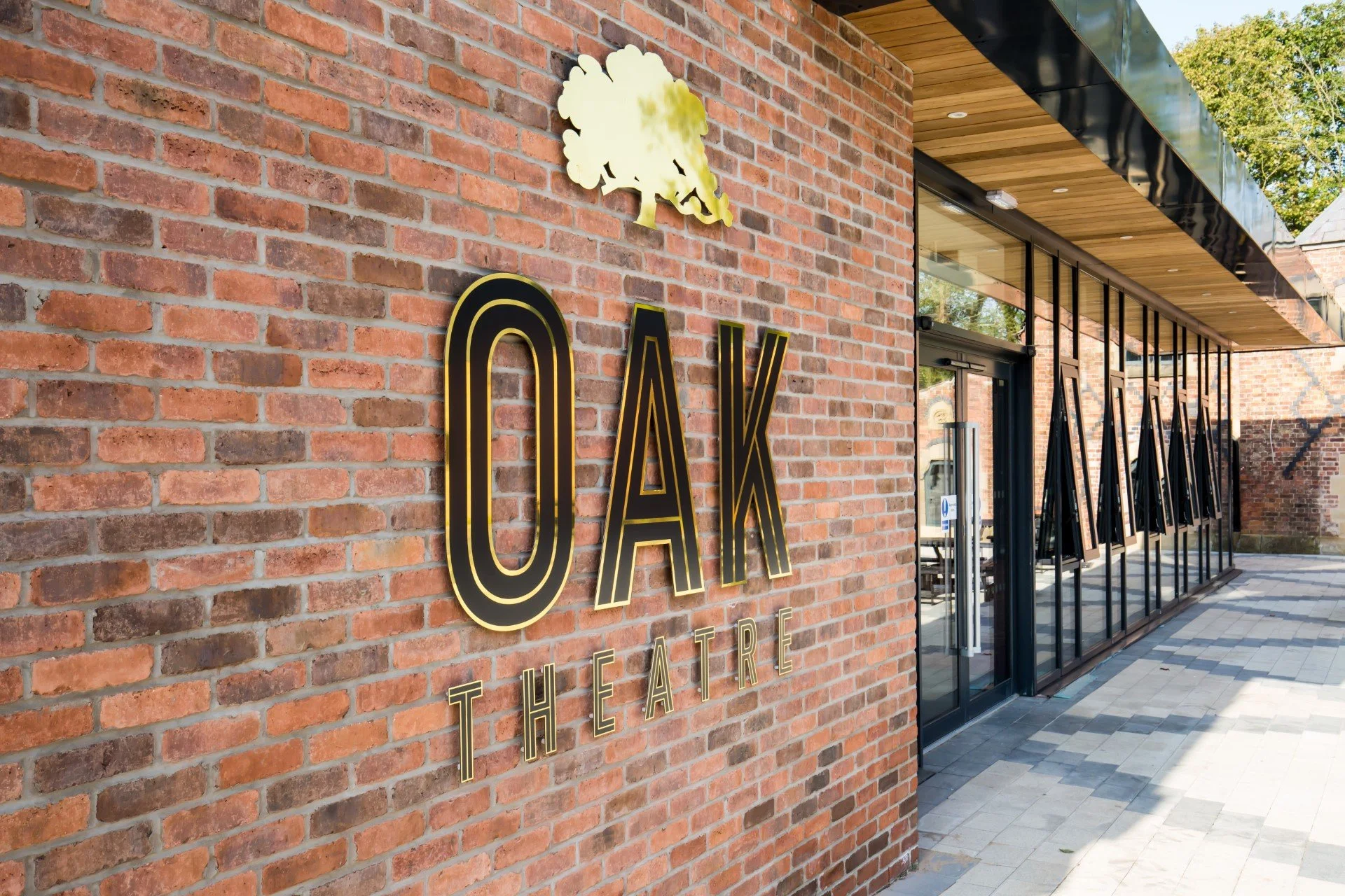 Exterior of a brick theater building with a yellow tree logo and large gold-lettered sign reading 'OAK THEATRE' on the wall.