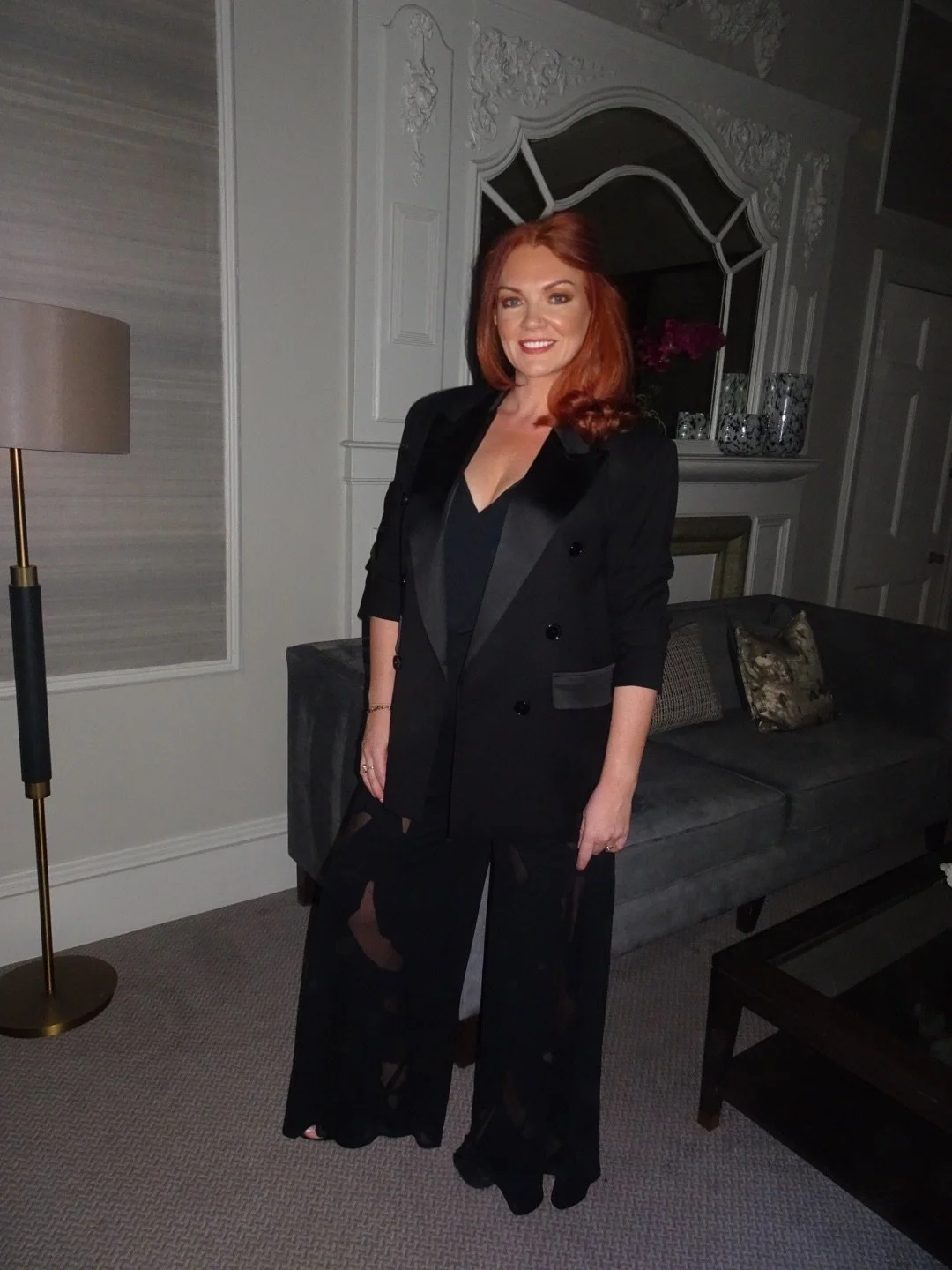 A woman with red hair wearing a black blazer, black top, and black sheer pants standing in a living room with gray sofa, decorative pillows, a mirror, and a floor lamp.