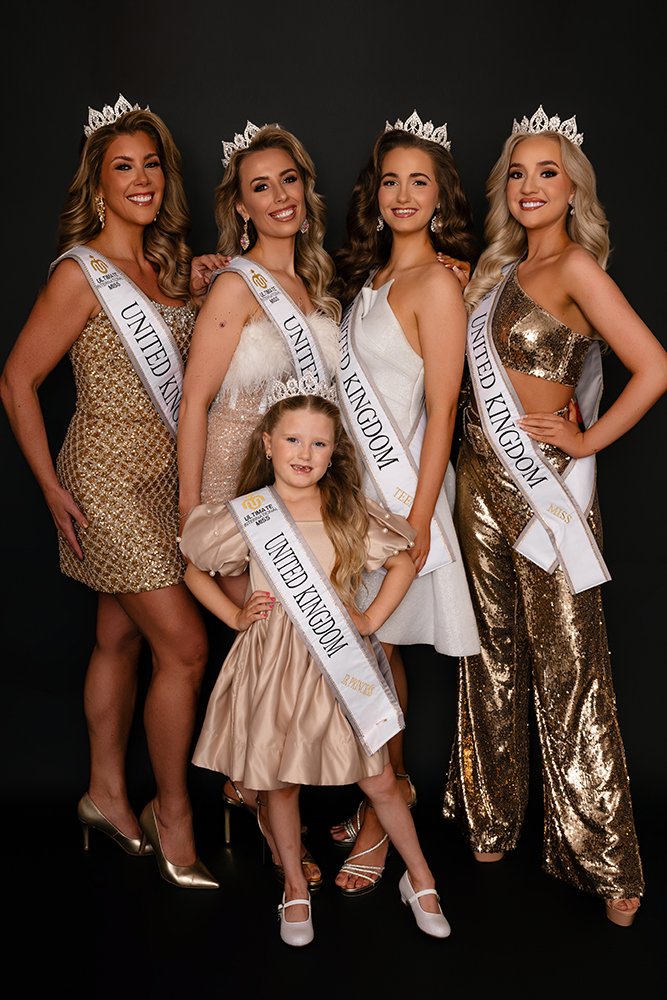 Group of five women and one young girl dressed in formal gowns, wearing crowns and sashes that read 'United Kingdom,' posing together against a black background, smiling for a photo, likely at a beauty pageant.