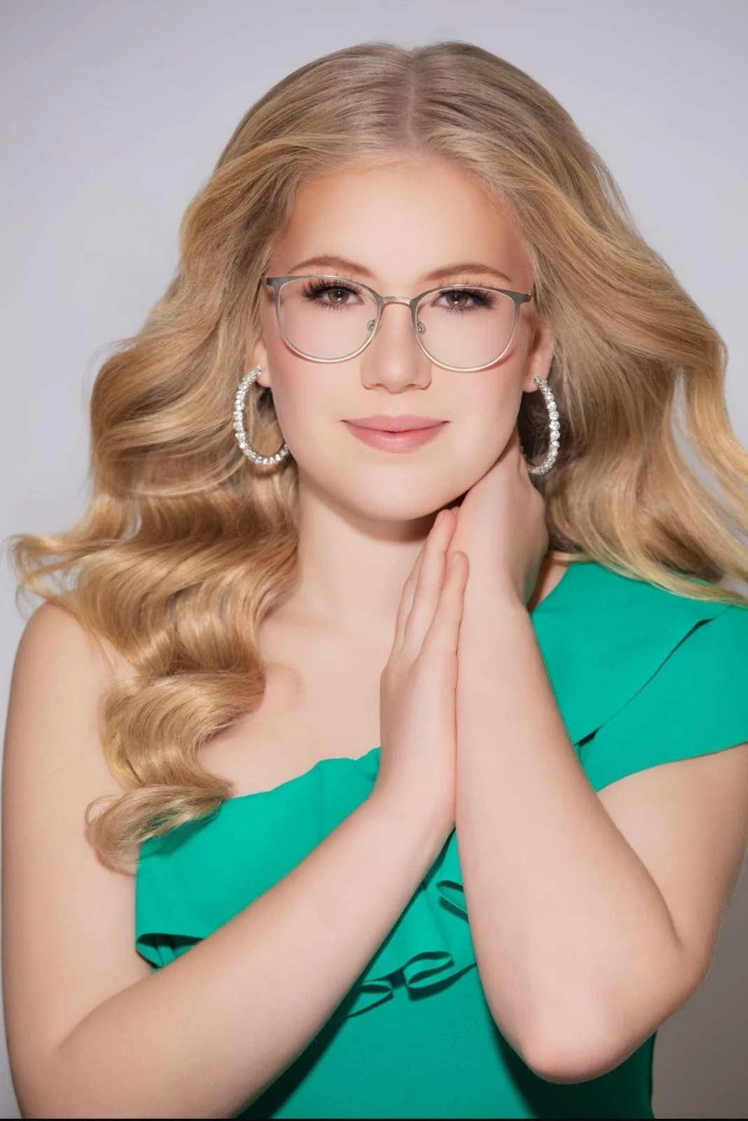 A young woman with wavy blonde hair, glasses, and hoop earrings, wearing a green dress and touching her face with her hands, smiling softly against a plain background.