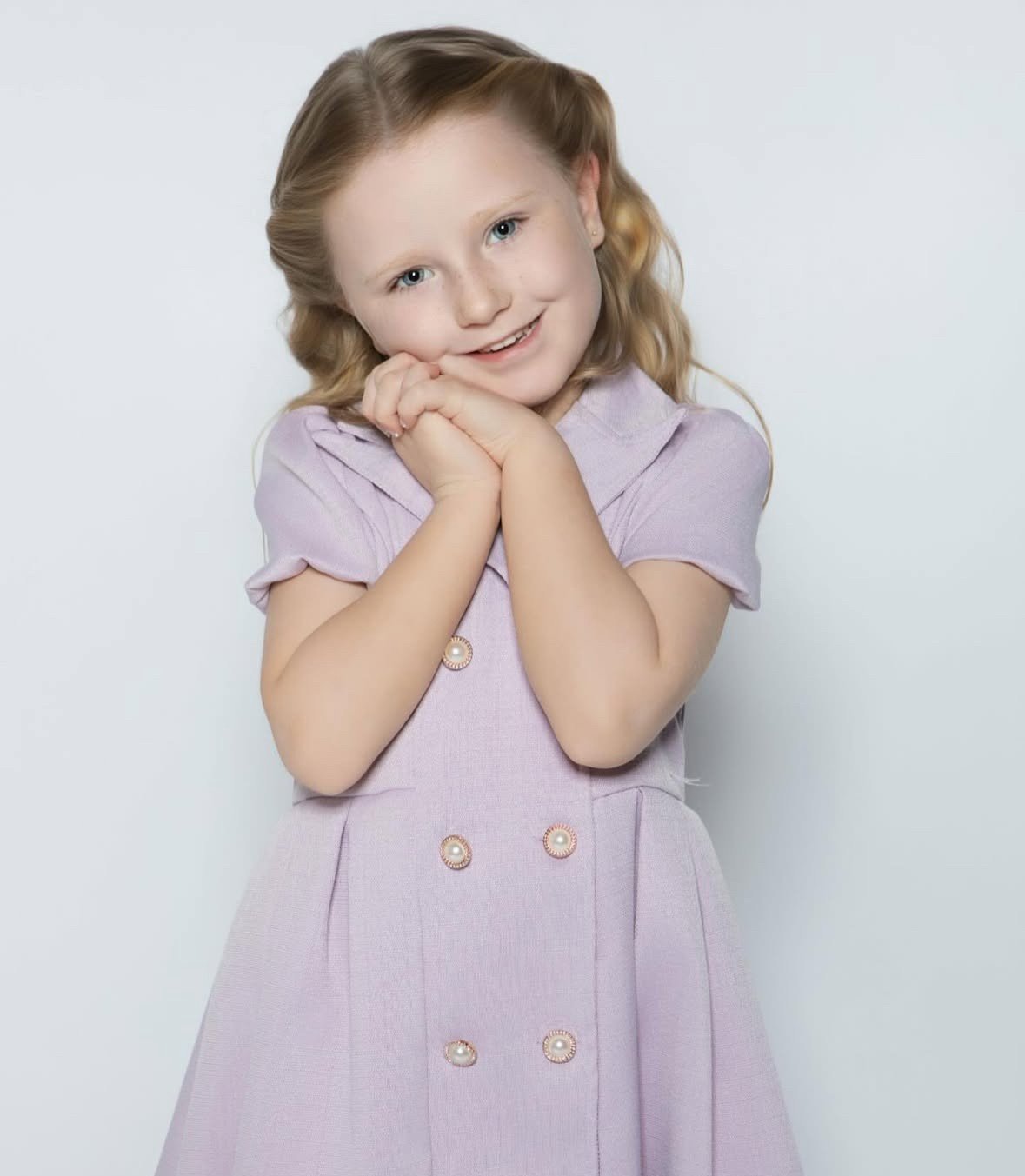 A young girl with blonde hair and blue eyes poses with her hands clasped under her chin. She is wearing a light purple dress with pearl embellishments and smiling at the camera against a plain light background.