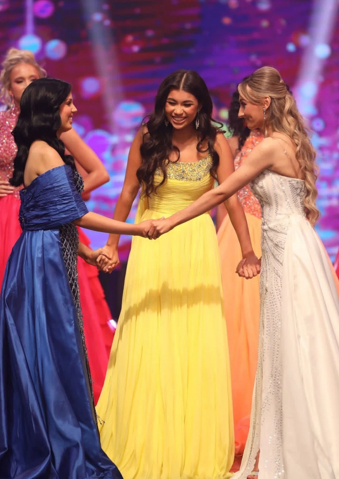 Group of women in colorful evening gowns holding hands on stage, celebrating at a pageant or formal event, with a vibrant, blurred background.