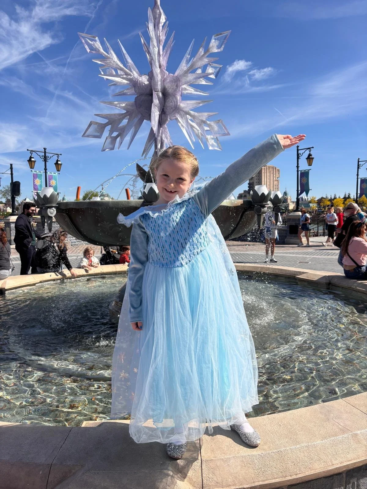 A girl dressed as Elsa from Disney's Frozen standing in front of a fountain with a snowflake sculpture, during a sunny day with a blue sky. People are sitting and walking around in the background.