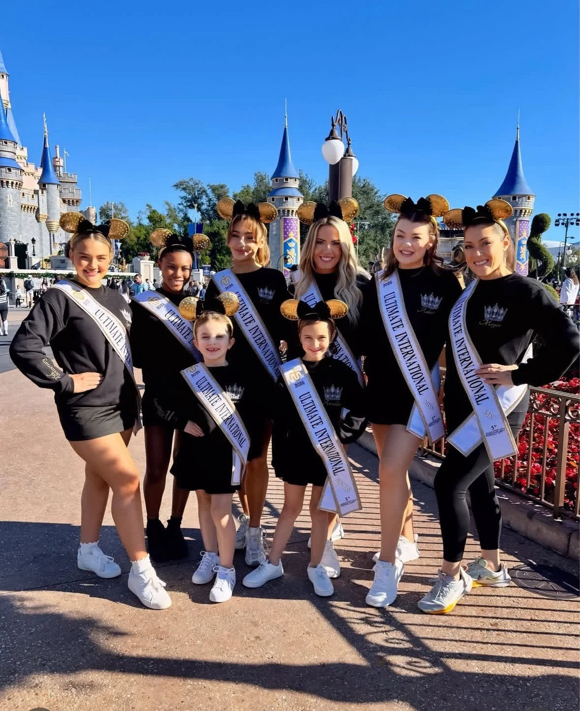 Group of young women and girls dressed in matching black outfits with Disney-themed Minnie Mouse ears, posing at Disneyland with Cinderella's castle in the background. They are wearing sashes that read 'Ultimate International'.