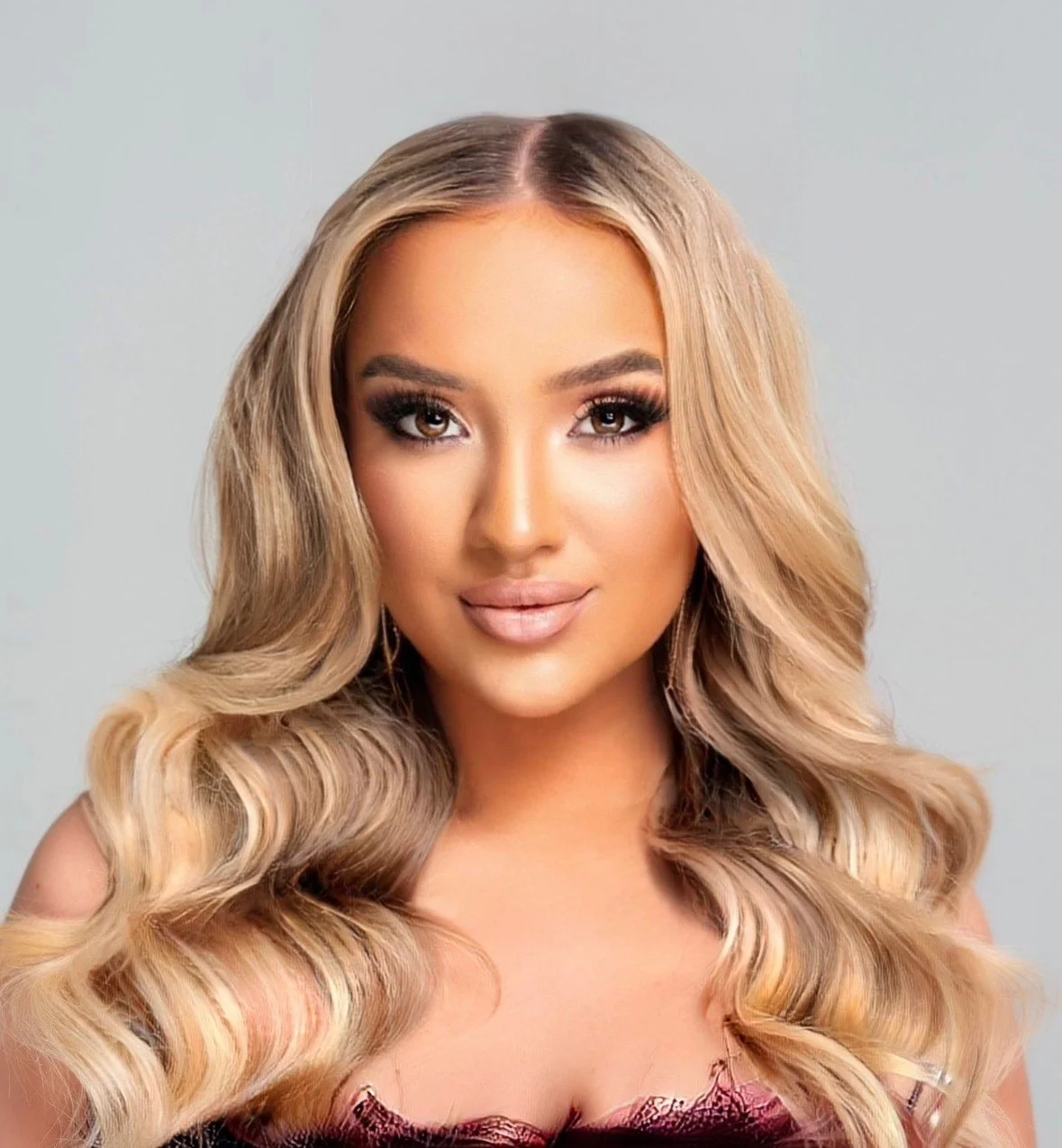 A woman with long, wavy blonde hair, makeup with smoky eyes, and neutral lipstick, wearing a dark red lace top, against a plain light background.