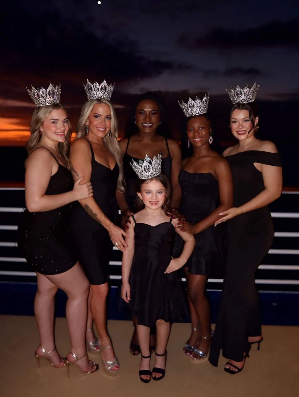 Six women and one girl wearing black dresses and crowns, standing on a boat deck at sunset with dark clouds and the ocean in the background.