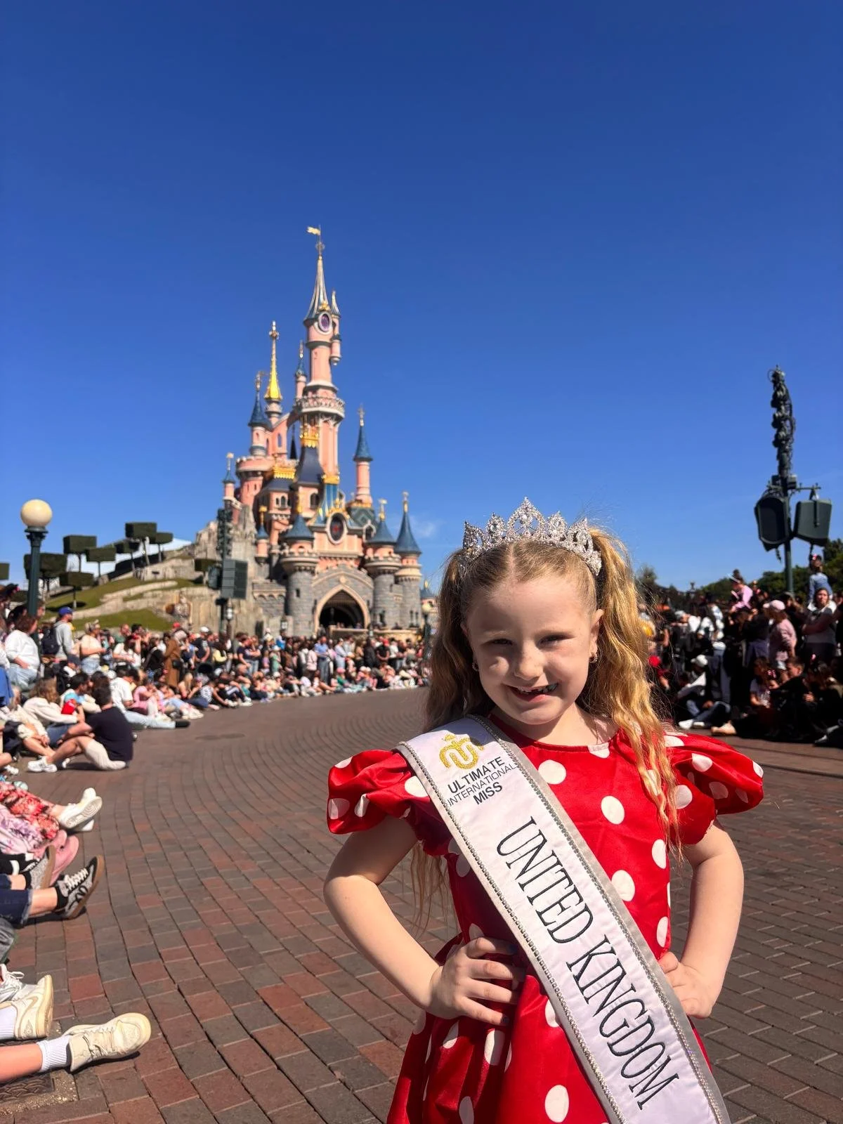 A young girl dressed as a princess with a crown, wearing a sash that reads "United Kingdom" and a tiara, standing in front of Sleeping Beauty Castle at Disneyland, with a crowd of people in the background.