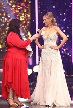 A woman in a red dress holding a microphone and an envelope, interviewing a woman in a white gown on stage during a formal event or award ceremony, with colorful lights and a starry background.