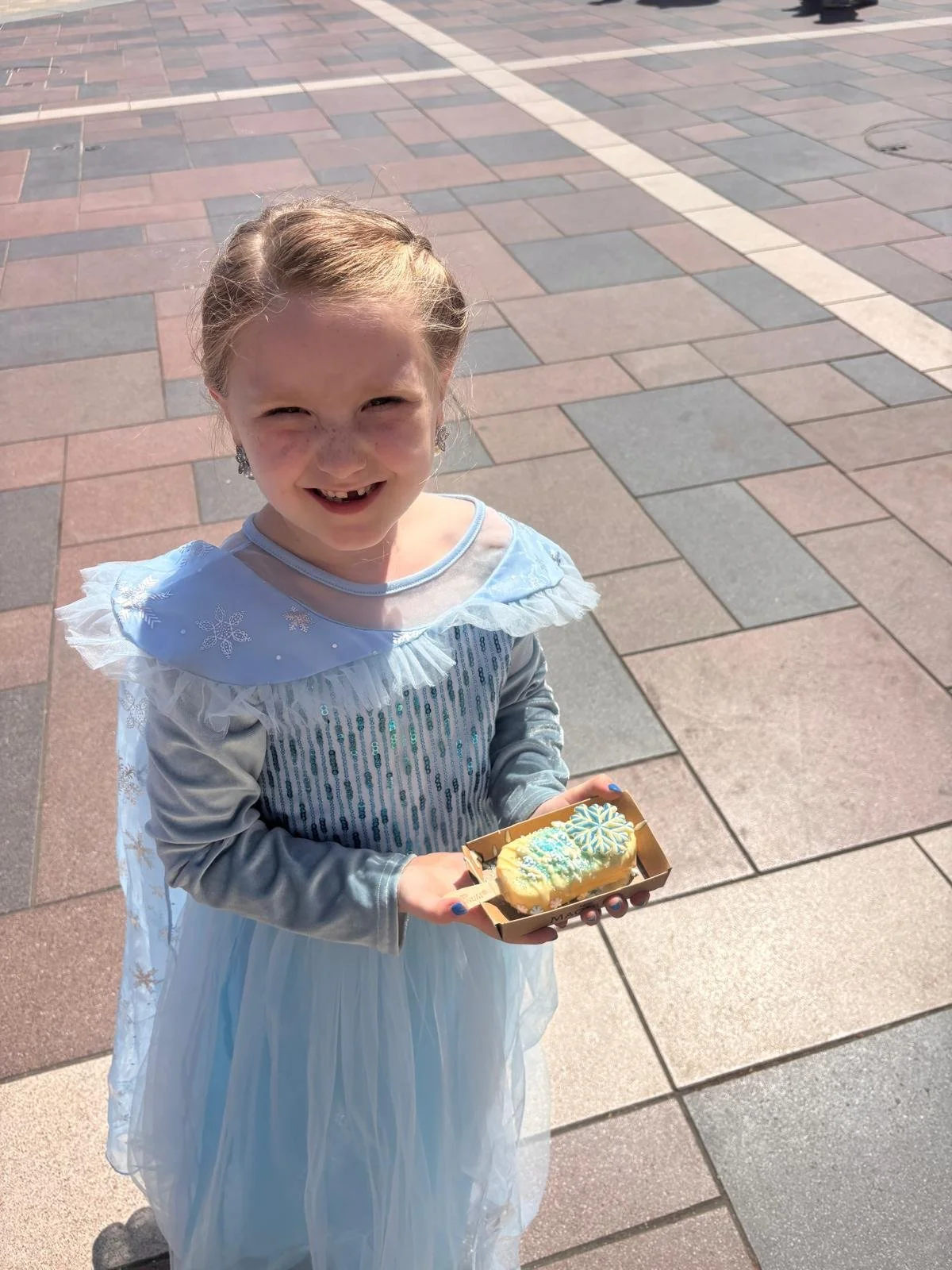 A young girl dressed as Elsa from Frozen is smiling and holding a small tray with a decorated cookie or pastry. She is outdoors on a brick-paved area, wearing a light blue dress with a sheer cape and sparkly details.