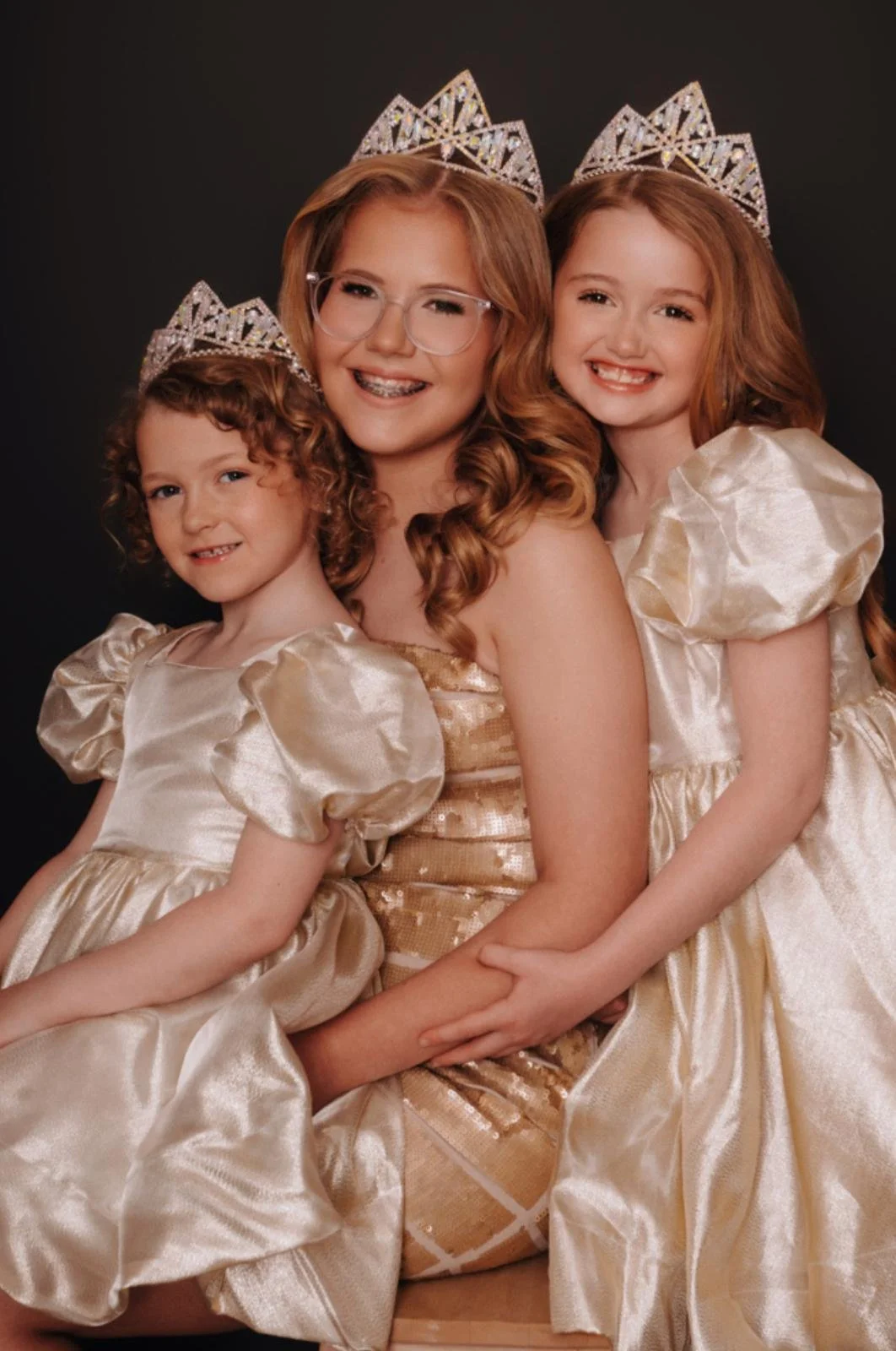 Three young girls and one woman, all wearing princess costumes with gold dresses and tiaras, posing together against a black background.