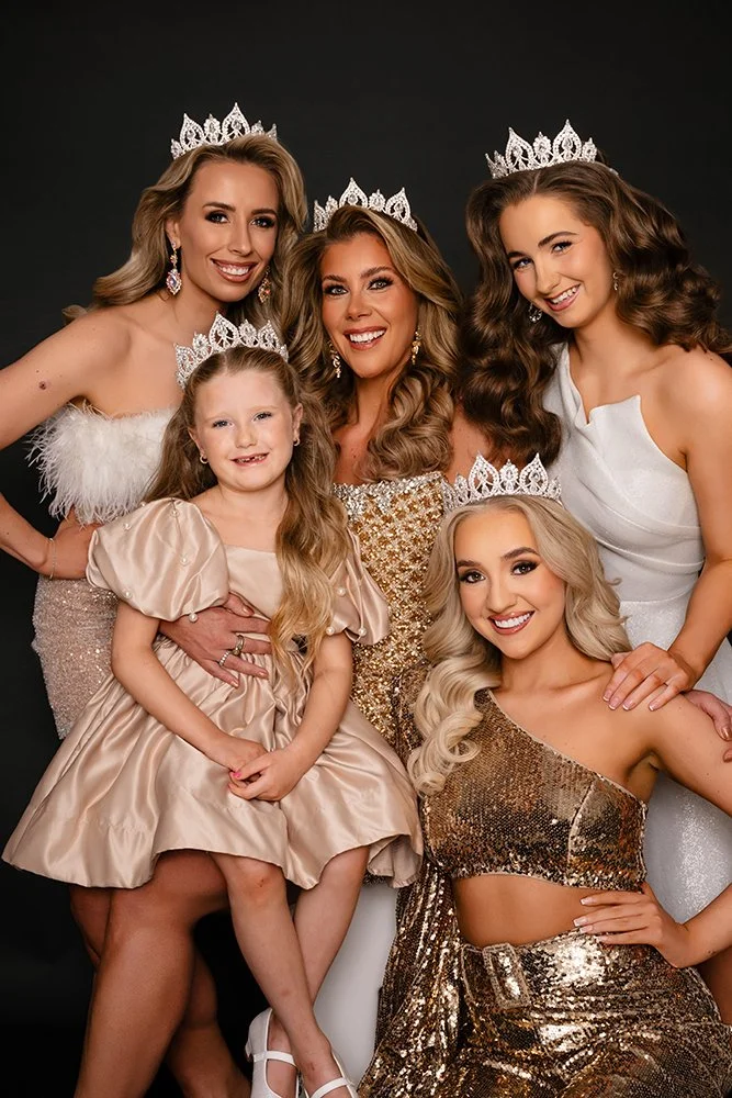 Five women and one young girl wearing evening gowns and tiaras, posing together for a photo against a black background.