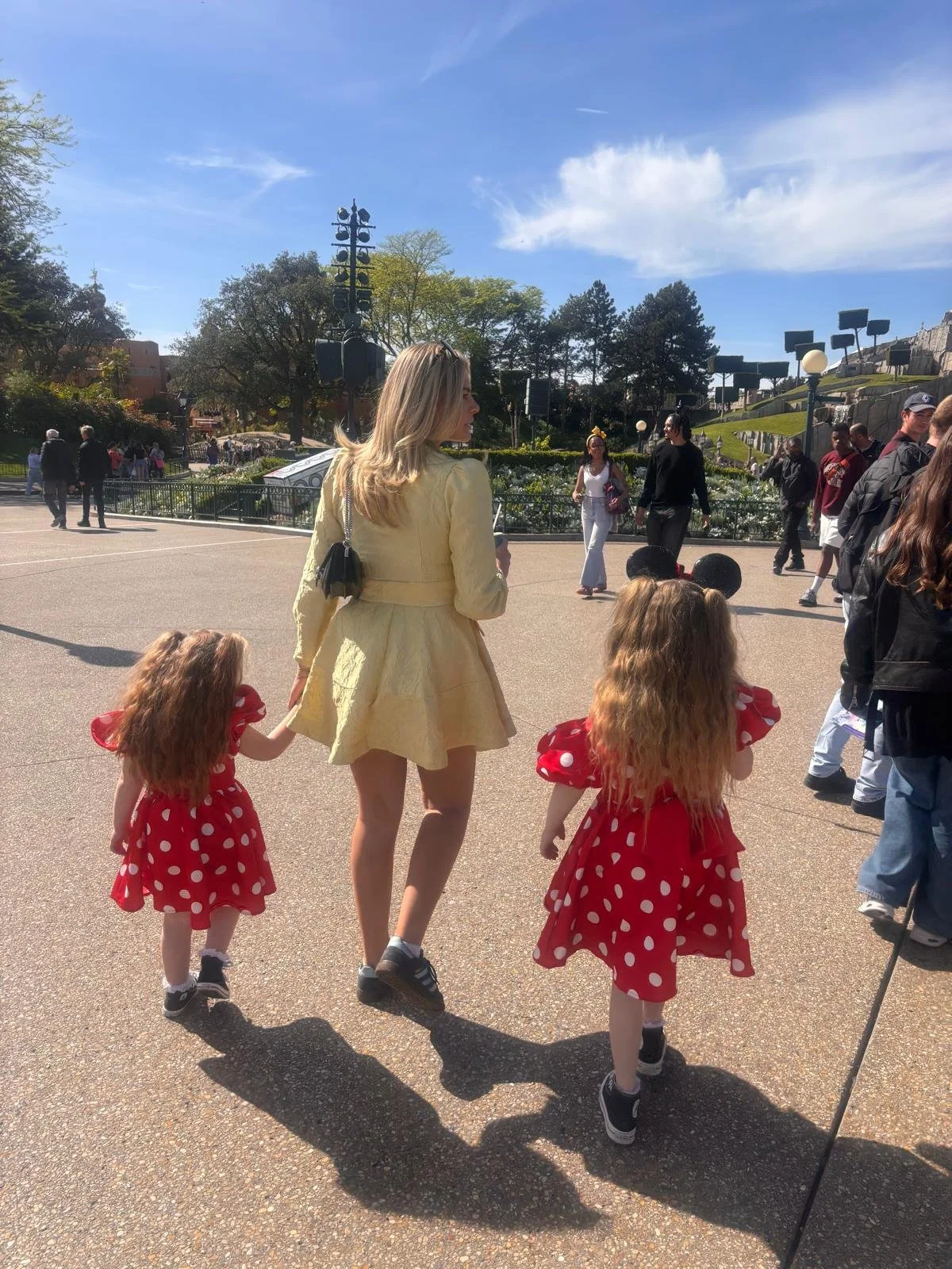 A woman with blonde hair in a yellow dress holding hands with two young girls in red polka dot dresses, walking outdoors on sunny day with a crowd of people around and park scenery in the background.
