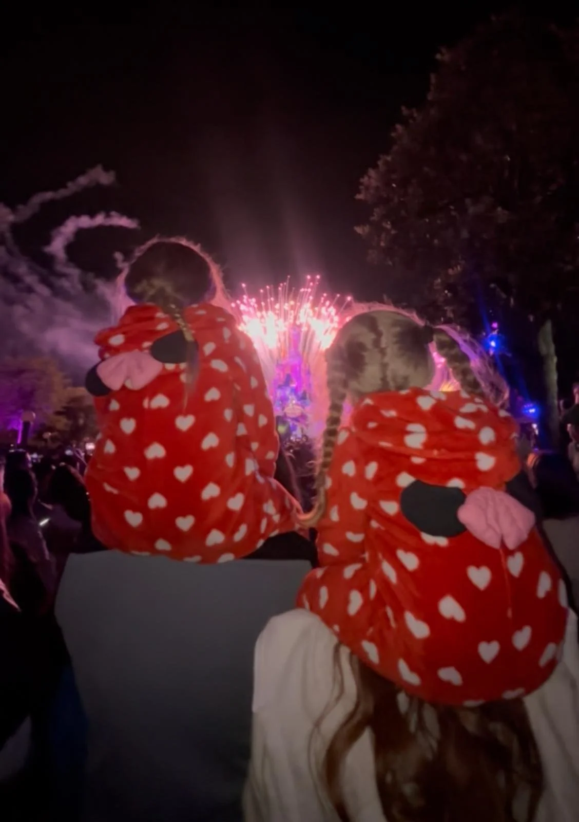 Two children wearing red hooded jackets with white hearts, sitting on an adult's lap, watching a fireworks display at night.