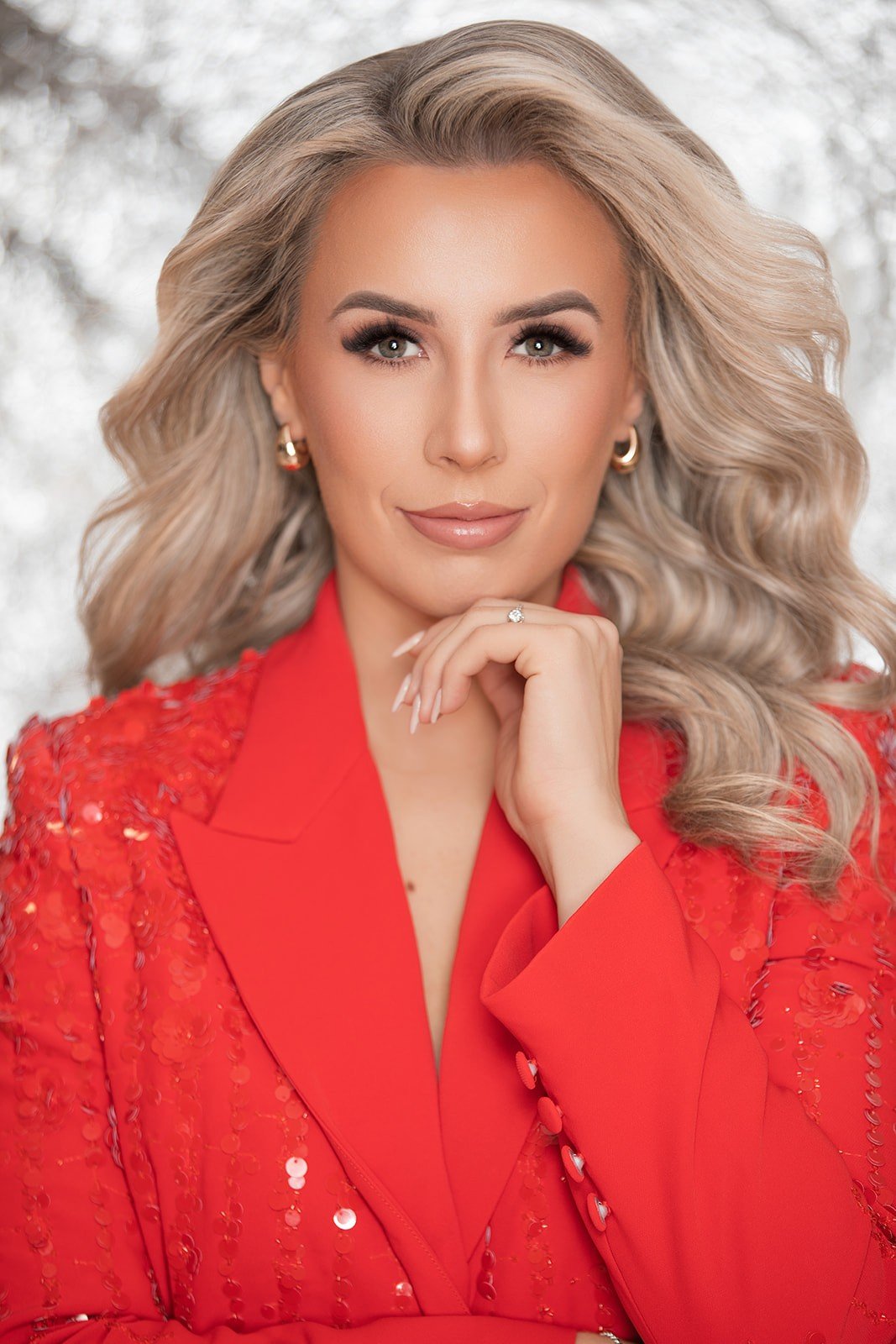 A woman with blonde, wavy hair and light makeup, wearing a red blazer with sequin embellishments and gold hoop earrings, poses with her hand near her chin in front of a blurred silver, sparkly background.