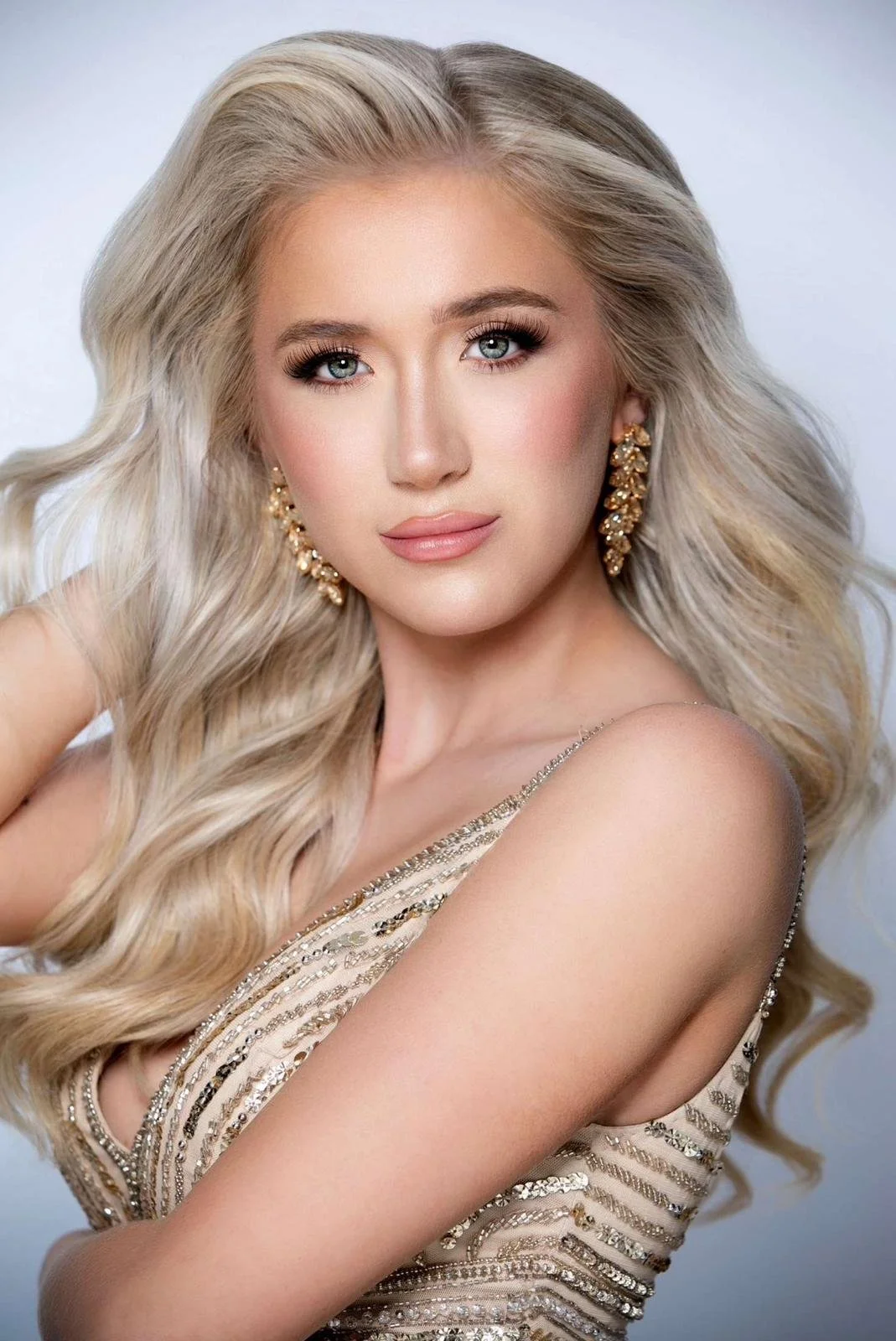 A young woman with long, wavy blonde hair and blue eyes wearing large gold earrings and a sparkling sleeveless dress, posing with her hand near her face.