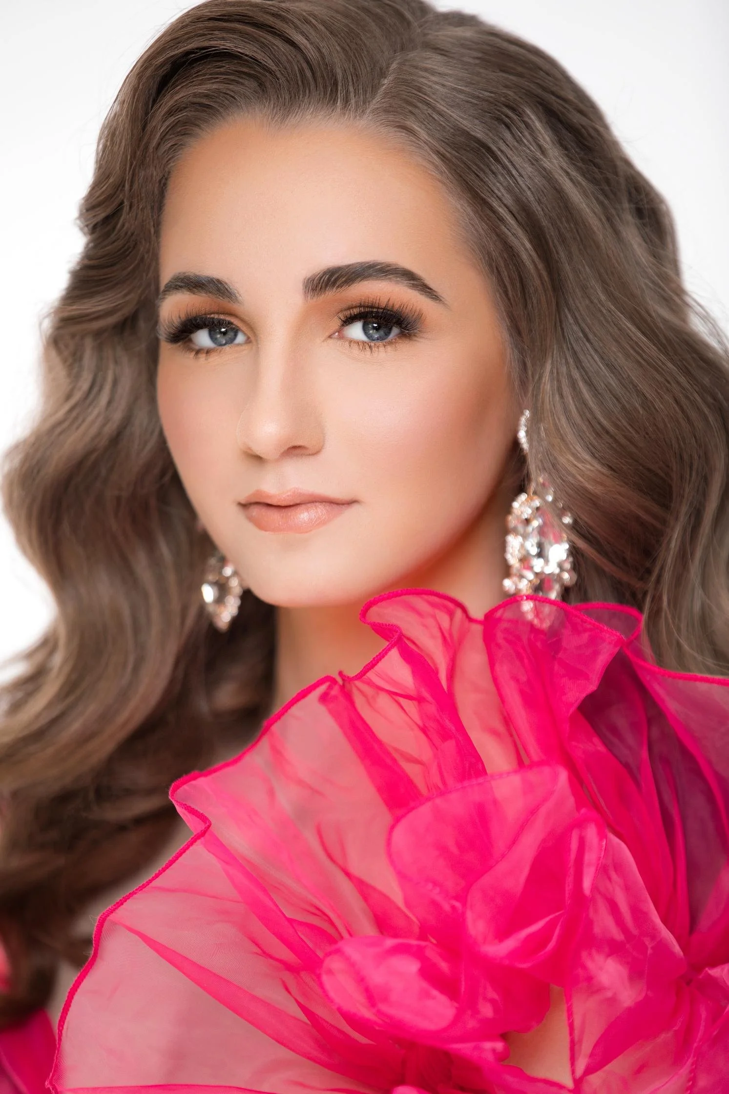 Close-up of a woman with long wavy brown hair, blue eyes, wearing sparkly earrings and a bright pink, ruffled, sheer dress against a plain light background.