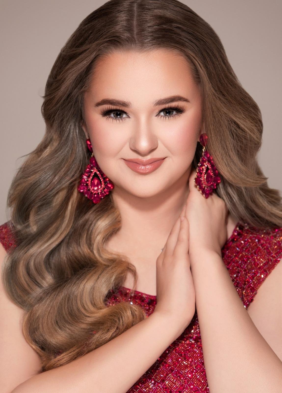A young woman with long, wavy blonde hair wearing pink earrings and a red, sparkly dress, smiling and posing with her hands clasped near her face.