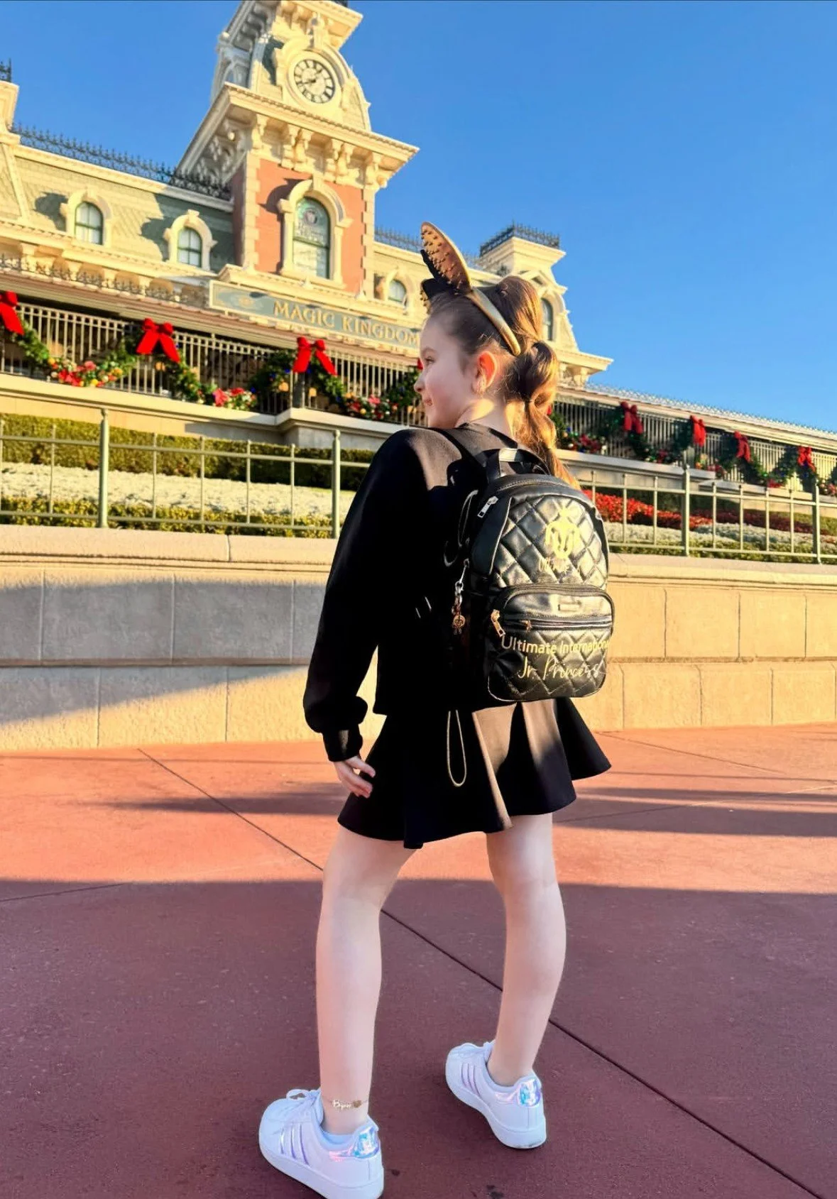 A young woman standing in front of Magic Kingdom at Disney World during daytime, wearing a black outfit, white sneakers, and a butterfly headband, with a black backpack.