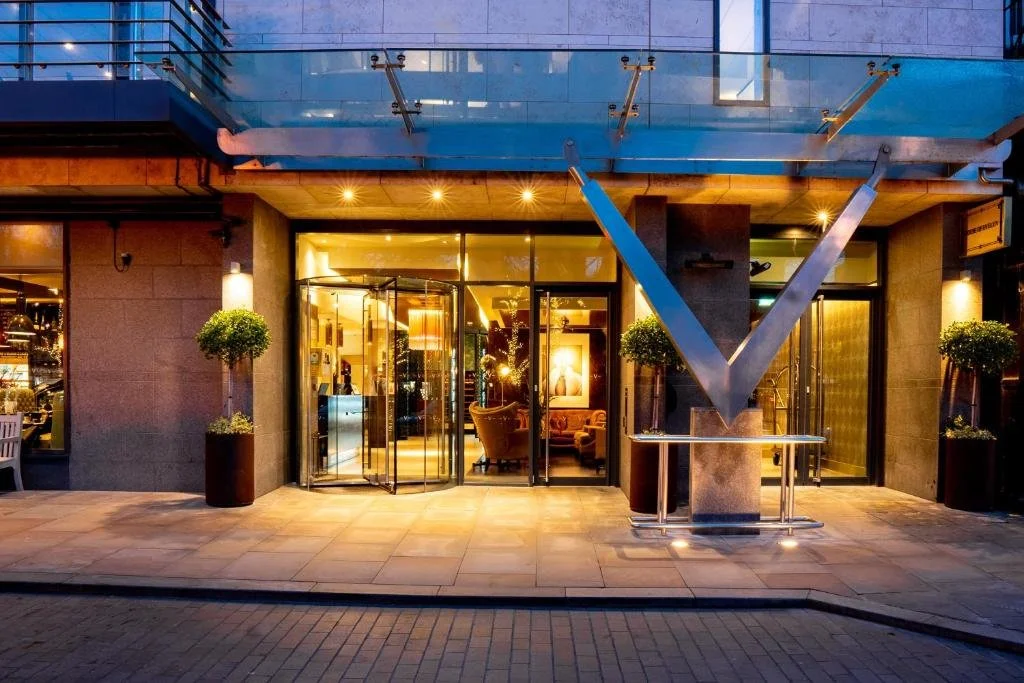 Hotel entrance with glass revolving door, lit warmly inside, two potted trees flanking the doorway, and a large metallic V-shaped structural element outside; the building has a modern glass and concrete facade, and the sidewalk is paved with light-colored tiles.