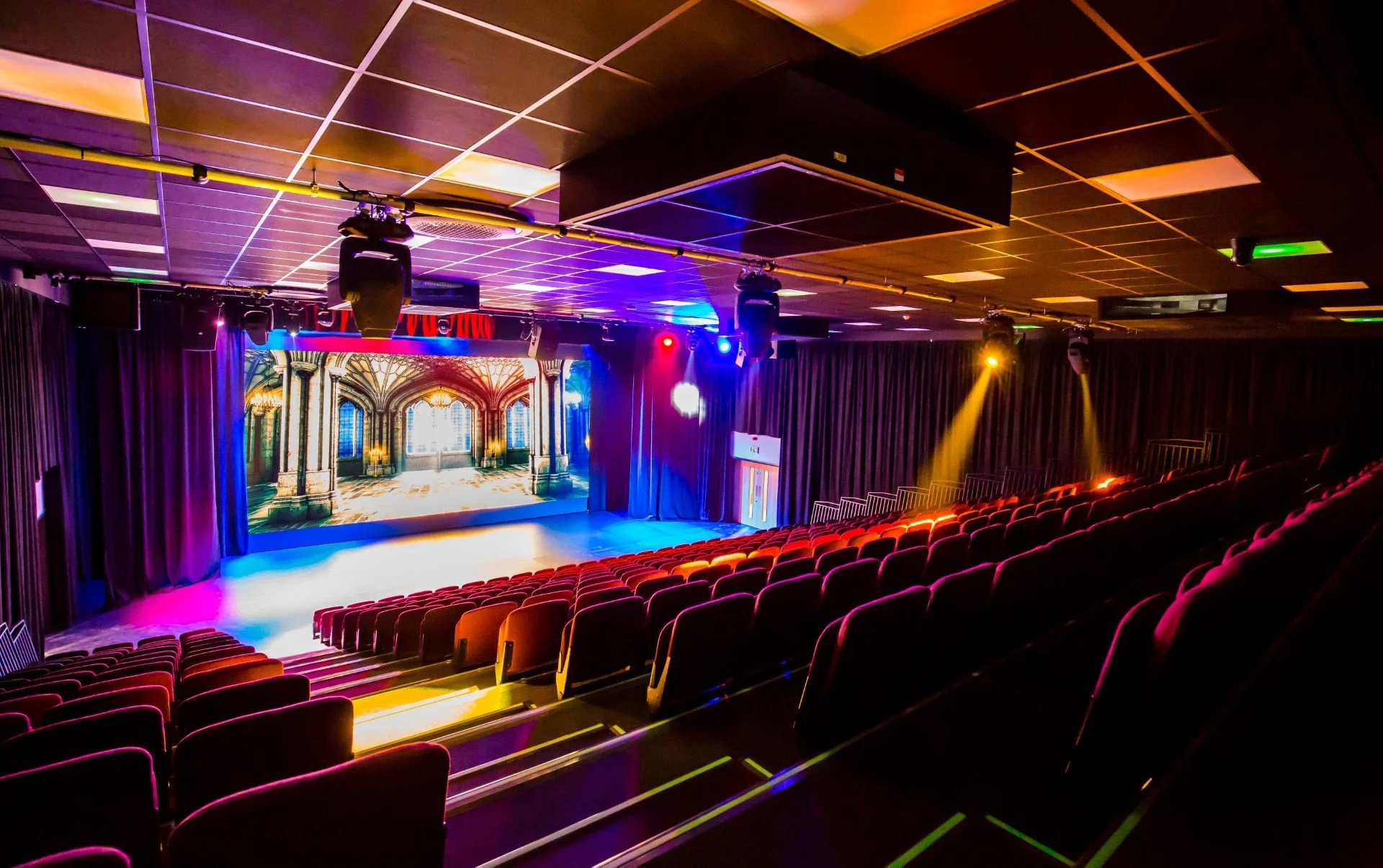 An empty theater with rows of red seats, a stage with a large screen displaying an ornate interior, and various colorful stage lights illuminating the space.
