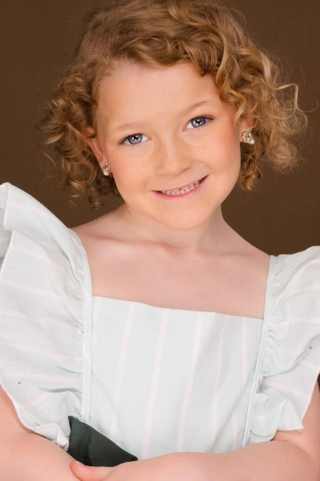 A young girl with curly red hair, blue eyes, and light skin, smiling at the camera. She is wearing pearl earrings and a white dress with puffed sleeves.