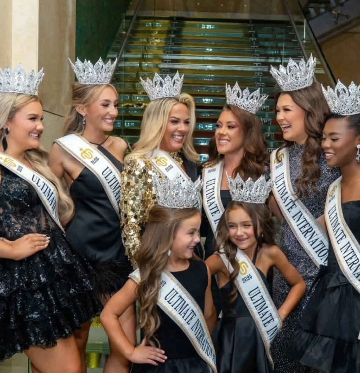 Group of beauty pageant contestants wearing crowns and sashes labeled 'Ultimate International 2023' and 'USA 2025,' smiling and talking with each other.