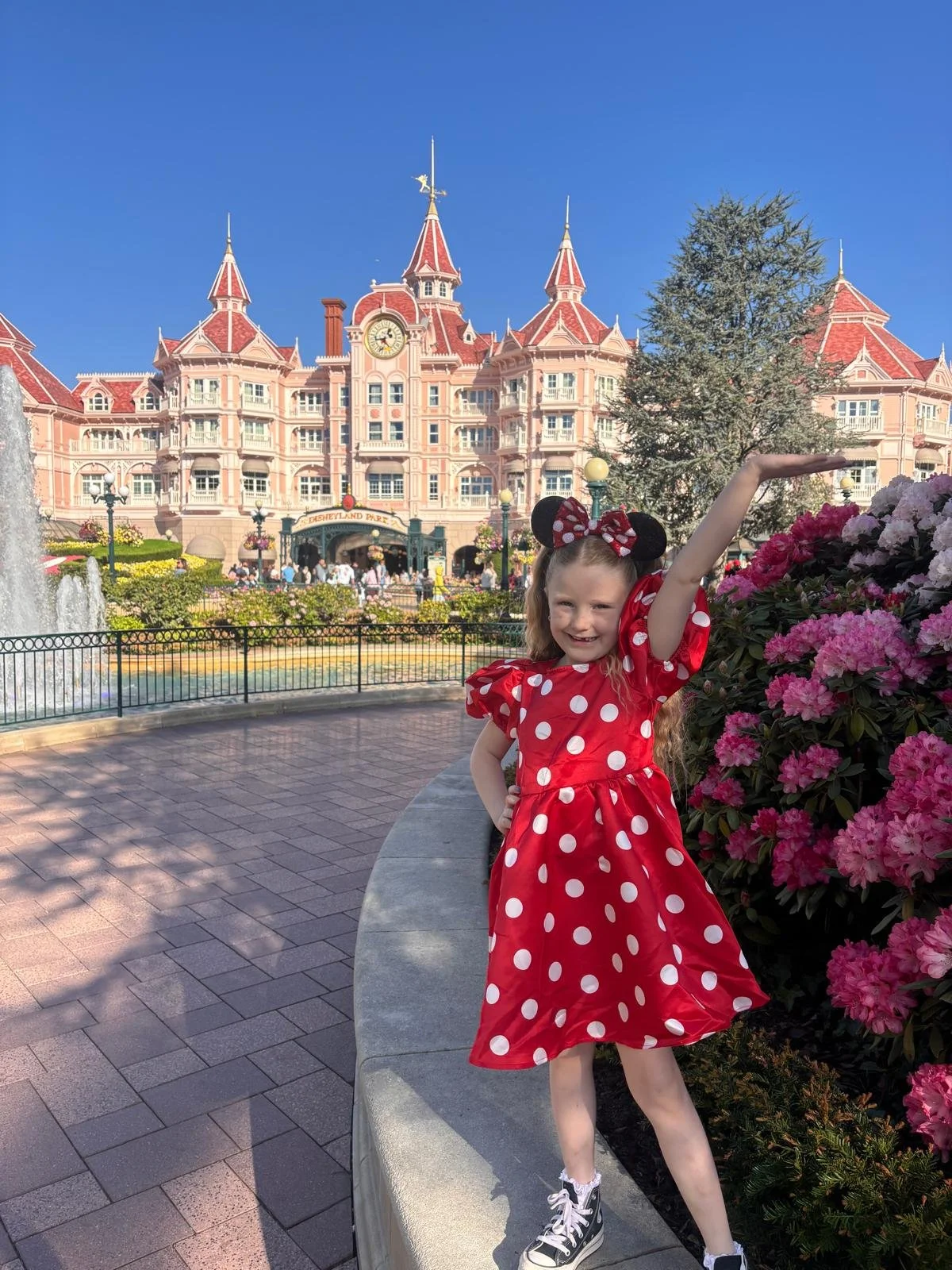 A young girl in Minnie Mouse ears and a red polka dot dress posing in front of the Disneyland Paris castle, with pink flowers and clear blue sky.