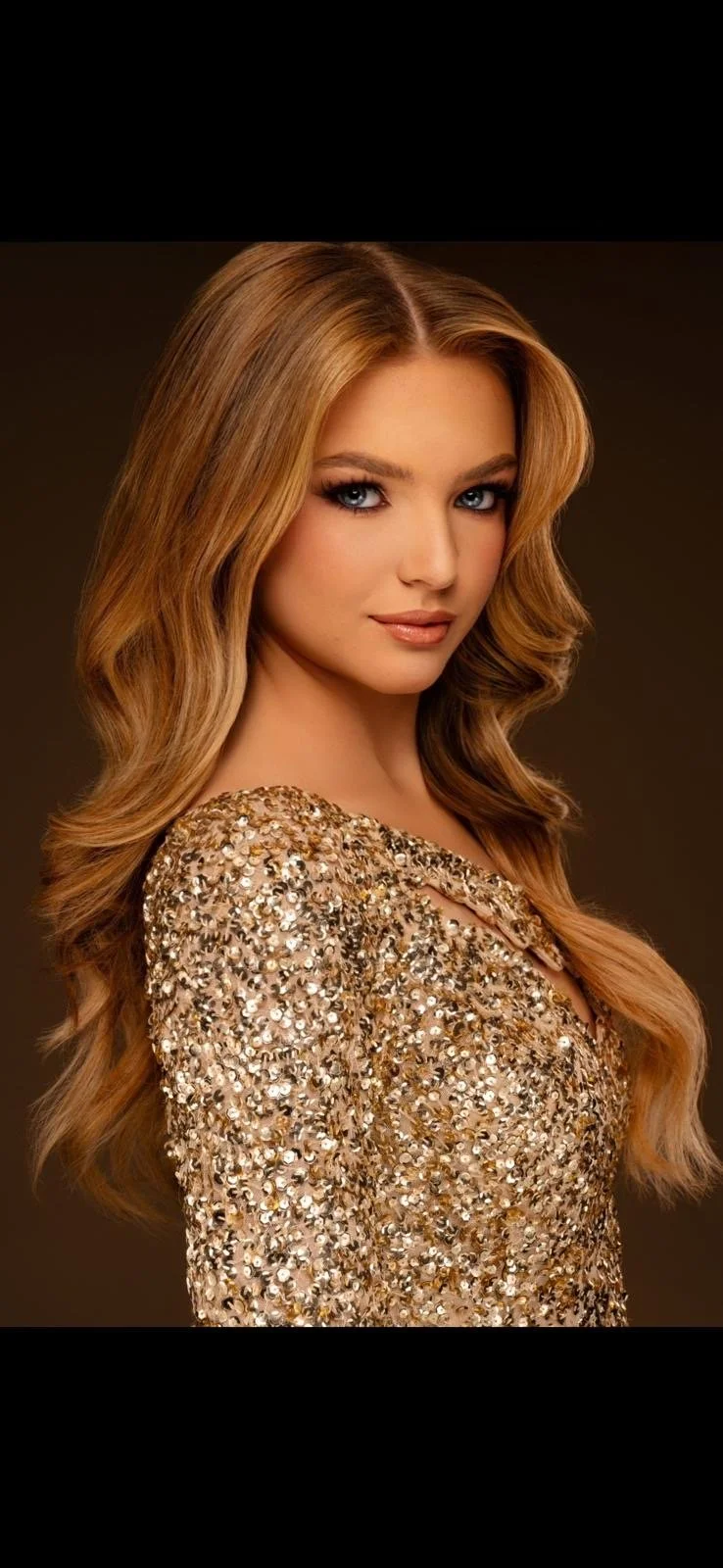 A woman with long, wavy blonde hair and bright blue eyes wearing a gold sequined dress.