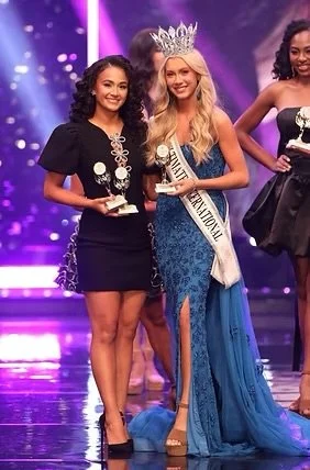 Beauty pageant winners on stage, with one wearing a crown and sash, and holding a trophy, celebrating their victory.