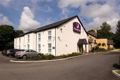 A white hotel building with a purple Premier Inn sign, parked cars, and a parking lot under a partly cloudy sky.