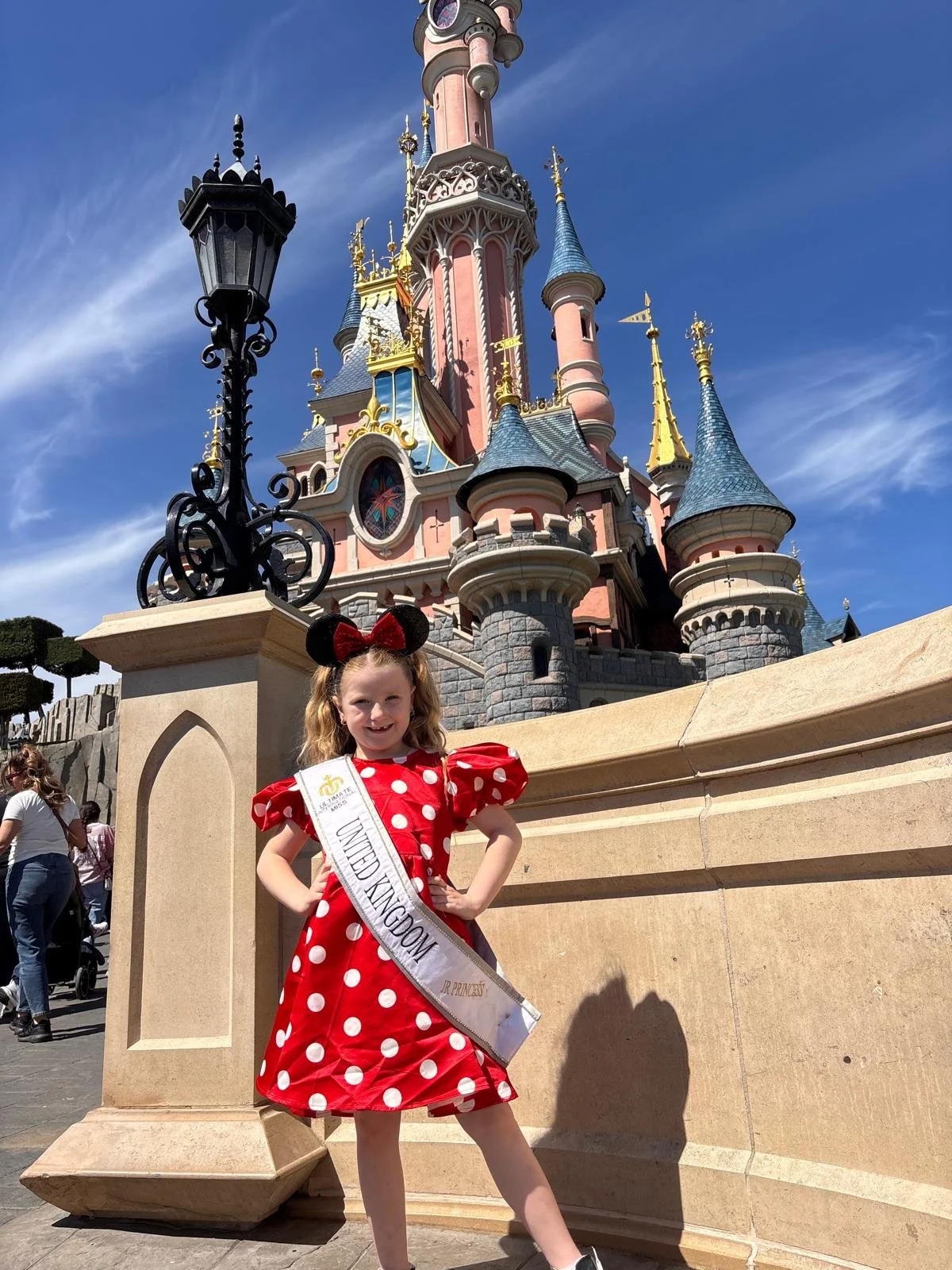 A woman dressed as Minnie Mouse wearing a red polka dot dress with a sash, standing in front of Sleeping Beauty Castle at Disneyland. The castle has pink walls, blue roofs, and gold accents. The sky is clear and blue, and there are a few other visito