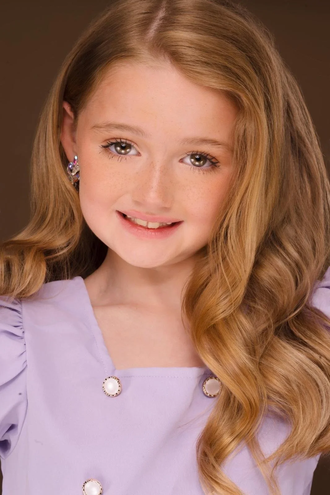 Young girl with long, wavy red hair and gray eyes, smiling softly, wearing a lavender top with pearl buttons and purple earrings.