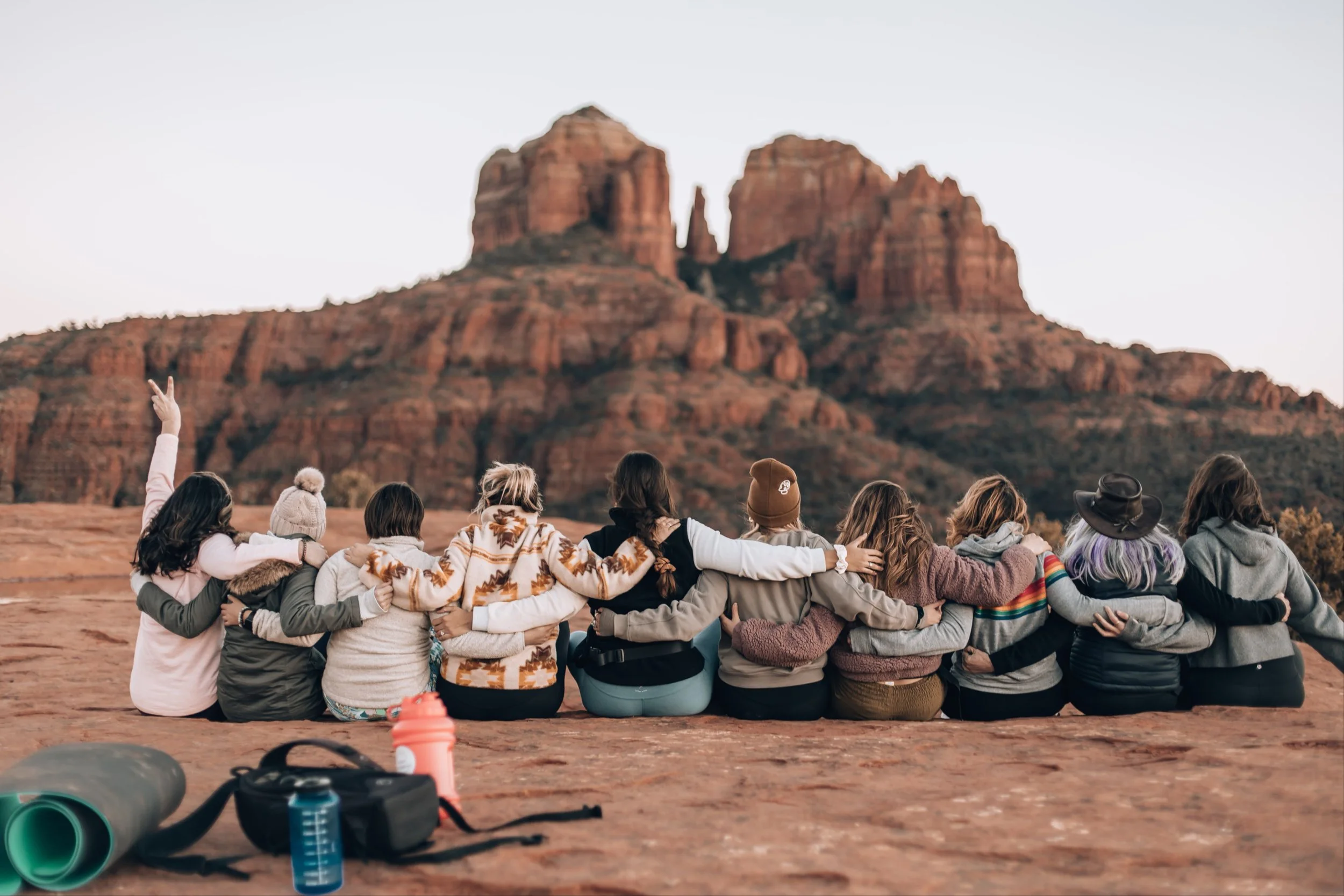 Group of friends sitting on rocky terrain with arms around each other, facing red rock formations at sunset in a desert landscape.