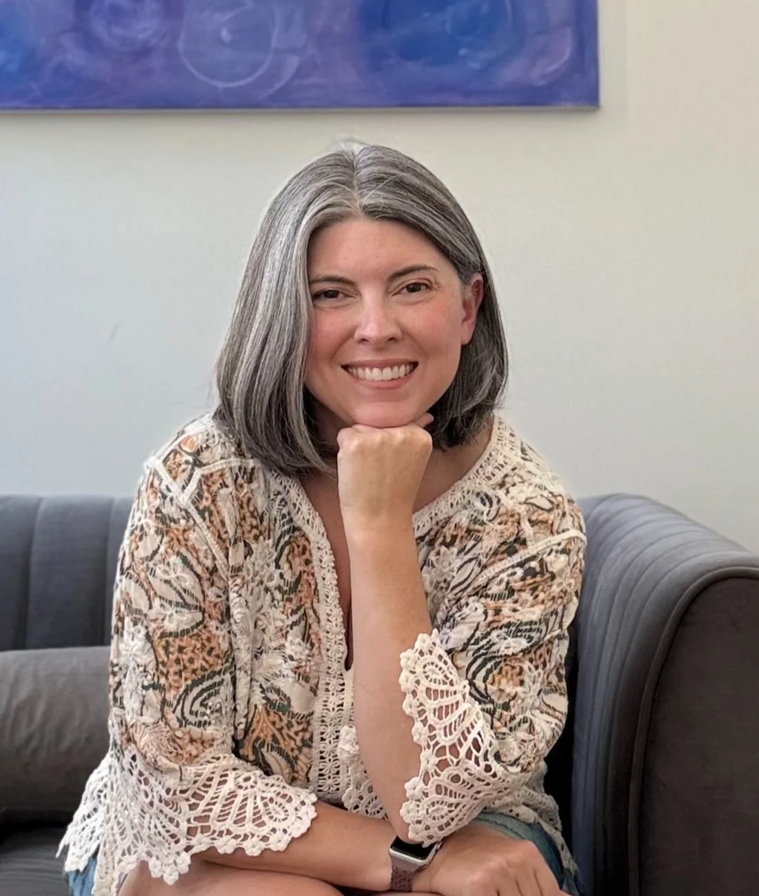 A woman with gray hair sitting on a sofa, resting her chin on her hand, smiling at the camera, in a room with a blue abstract painting.