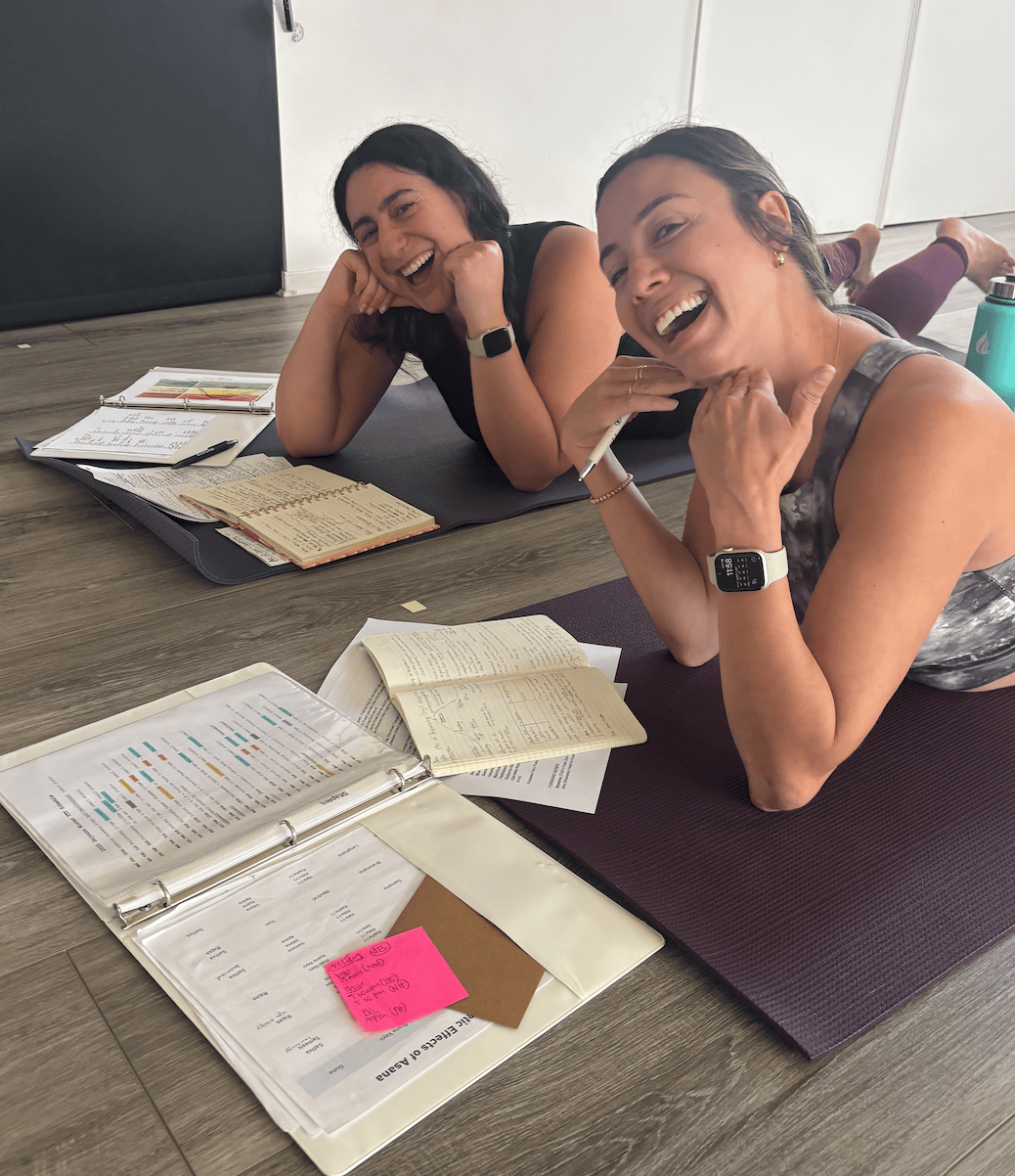 Two women lying on yoga mats on a wooden floor, smiling and looking at the camera, surrounded by notebooks, papers, and a water bottle, in a room with a dark door and white walls.
