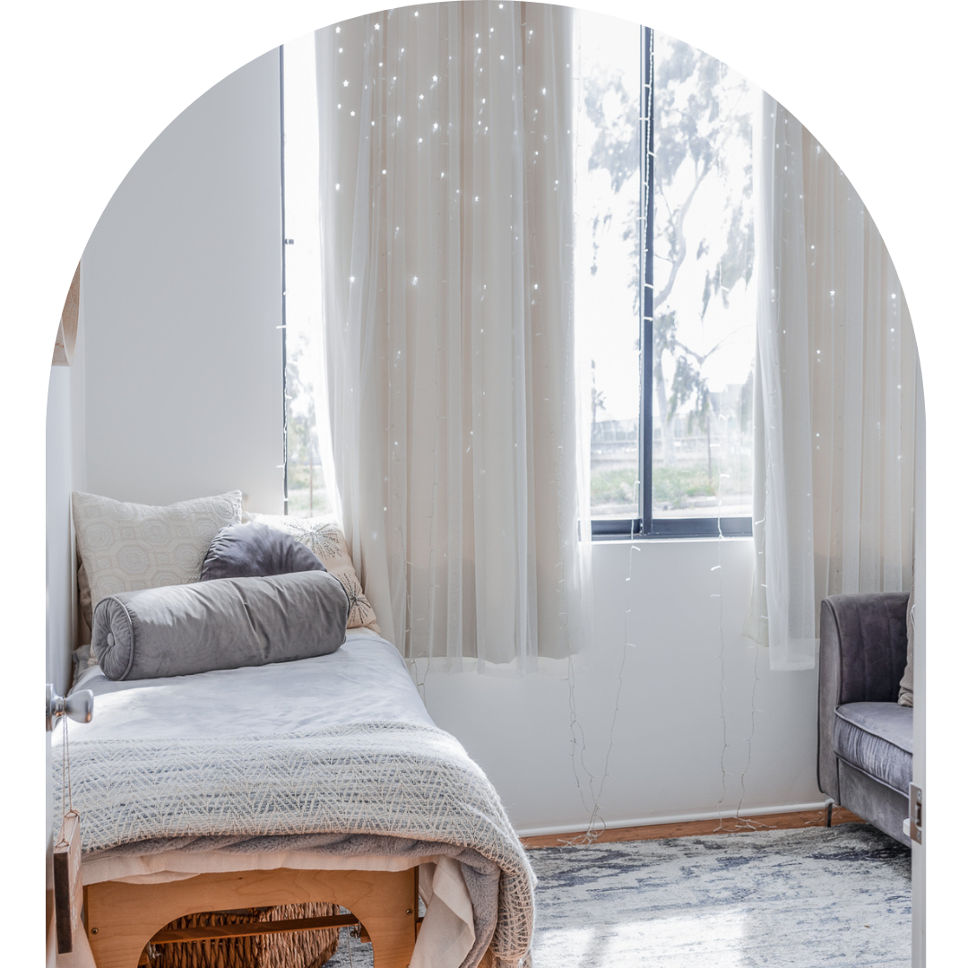 A bright bedroom with a bed on the left and a window with white curtains, fairy lights hanging in front of the window, and a gray armchair on the right.