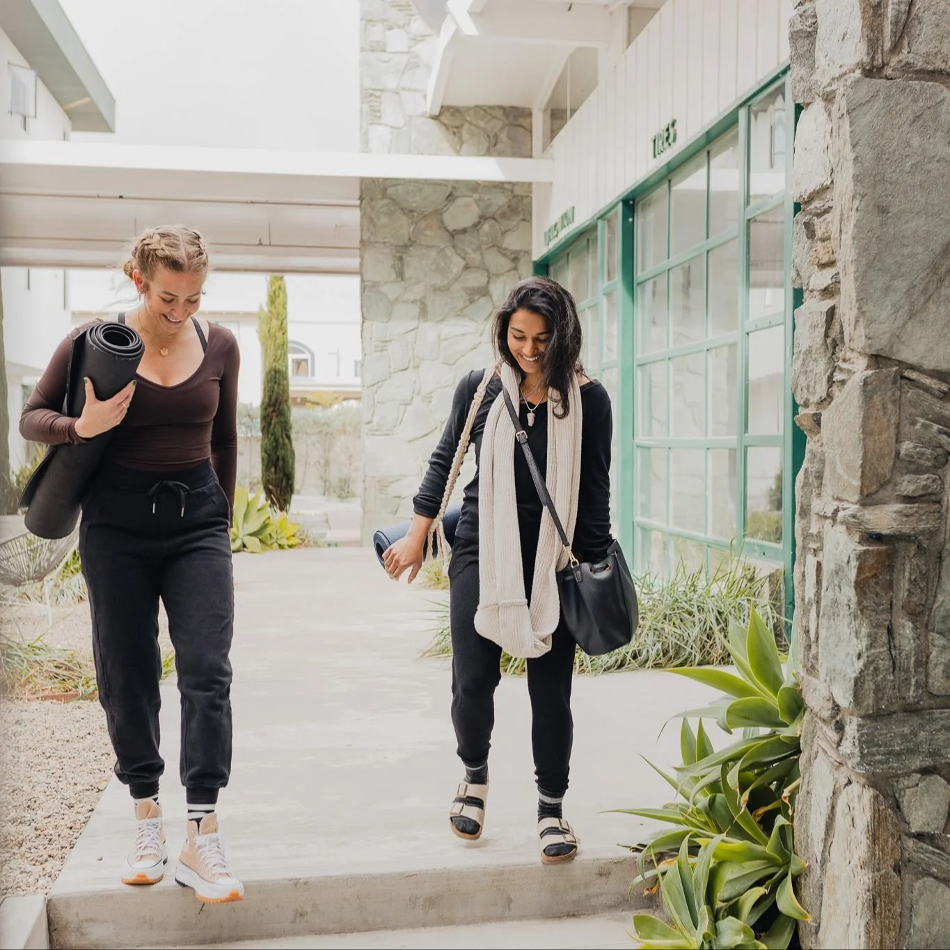 Two women walking outdoors, carrying yoga mats and wearing casual workout clothes, smiling and talking.