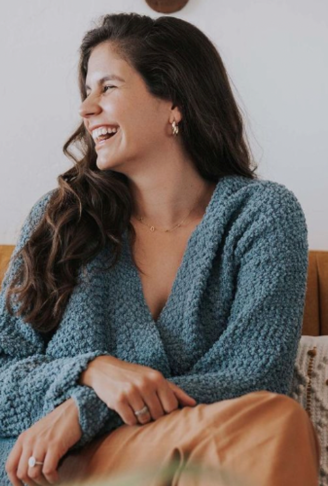 A woman with long, brown, wavy hair laughing and looking to her left, wearing a blue textured sweater, sitting on a wooden surface with a white wall in the background.