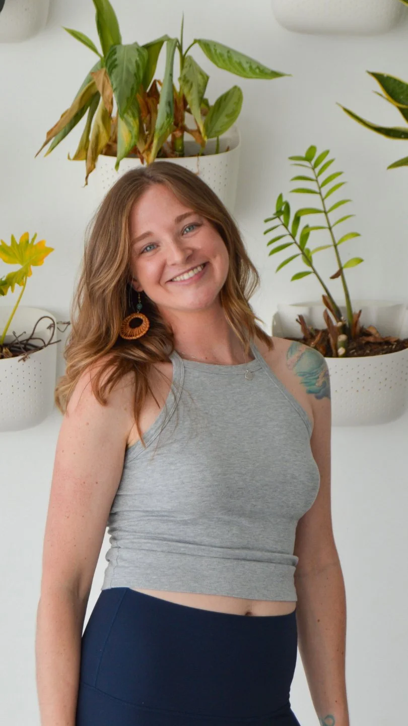 A woman with wavy light brown hair and earrings smiling in front of a wall with white planters filled with green plants.
