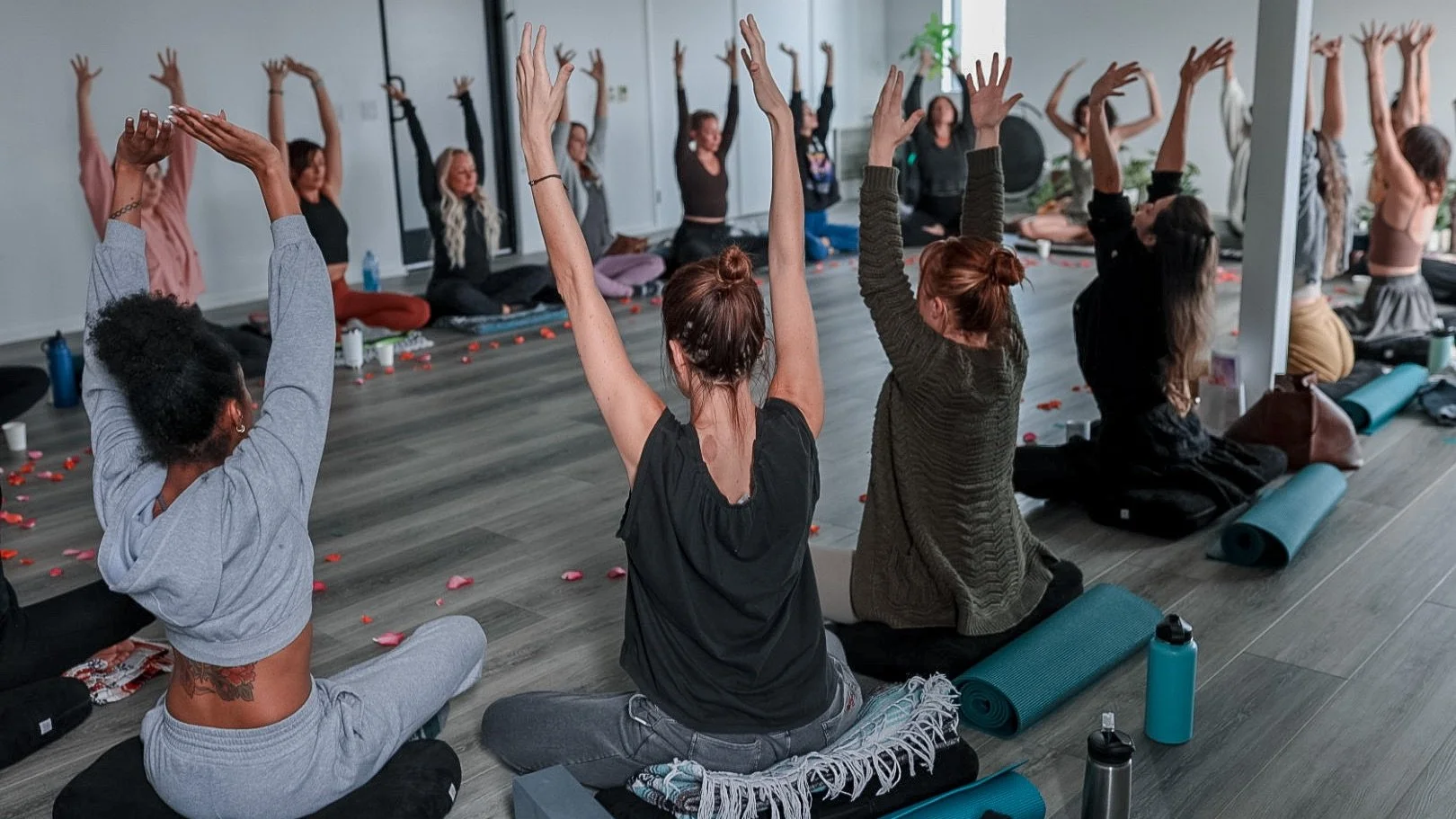 A group of people practicing yoga in a studio, sitting cross-legged or kneeling on mats with hands raised above their heads.