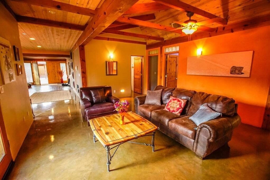 Living room with a brown leather sofa, blue and pink throw pillows, a wooden coffee table with a vase of flowers, and warm yellow walls with artwork, illuminated by ceiling and wall lights, and a doorway leading to other rooms.