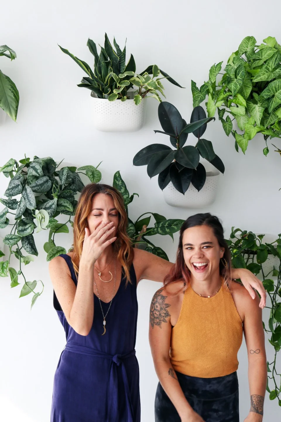 Two women laughing and smiling, one with her arm around the other's shoulder, against a background of green potted plants on a white wall.